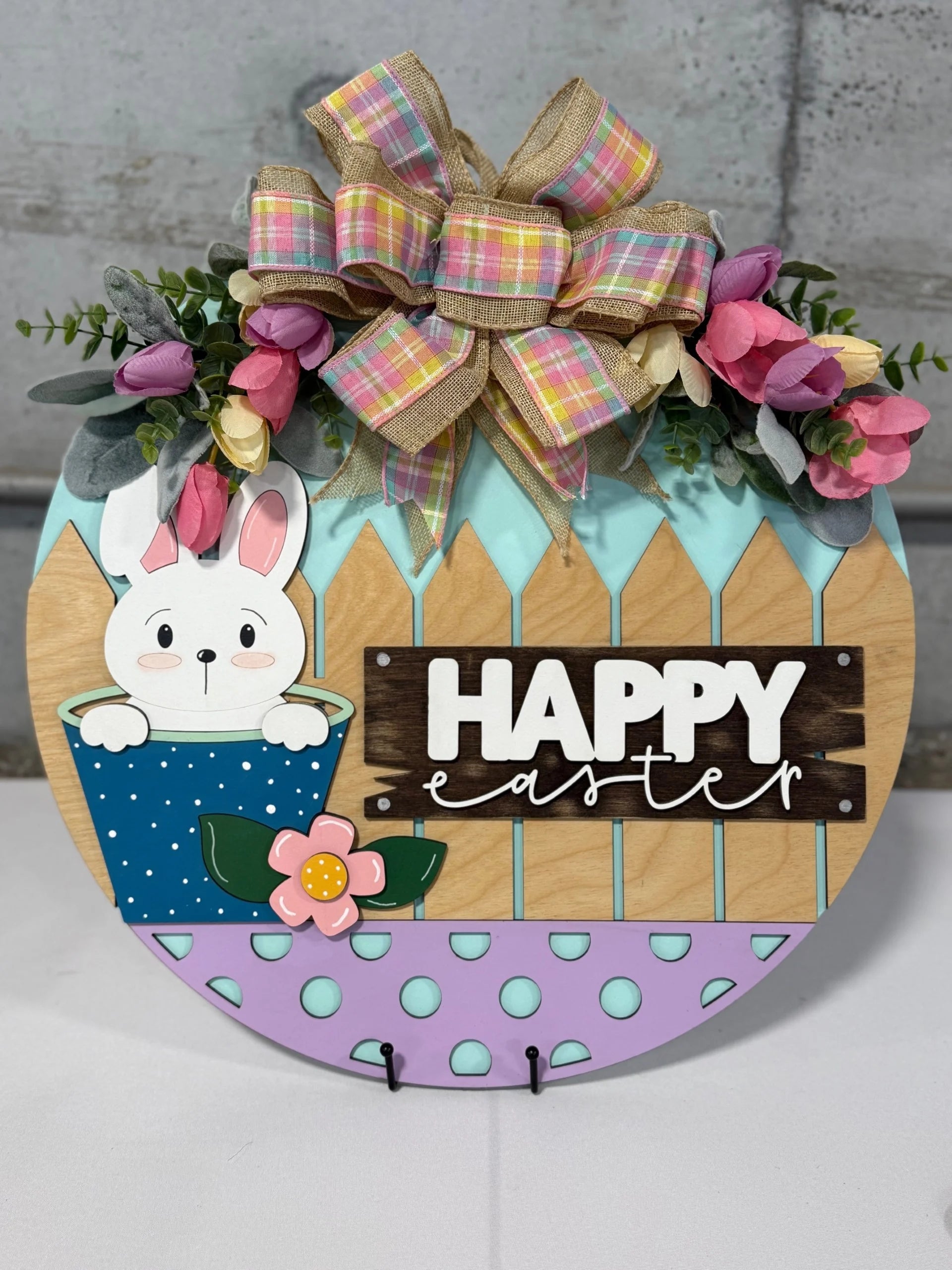 A decorative Easter sign features a cute white bunny peeking out of a blue polka-dotted pot. Above is a pastel plaid bow with greenery and pink tulips. The background resembles a wooden fence, and the words Happy Easter are written in white on a brown plank.
