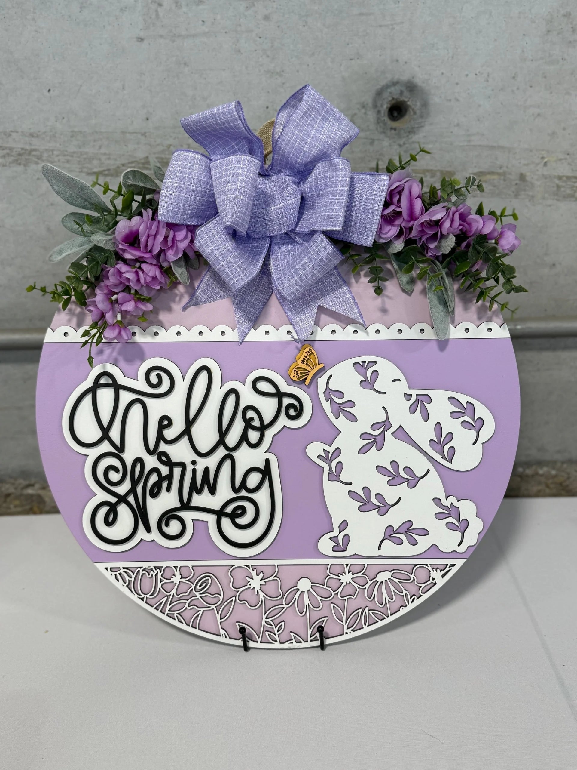 A round sign with a lavender and white design features the words Hello Spring and a decorative rabbit silhouette with floral patterns. Its adorned with a purple bow and greenery with purple flowers, set against a gray concrete background.
