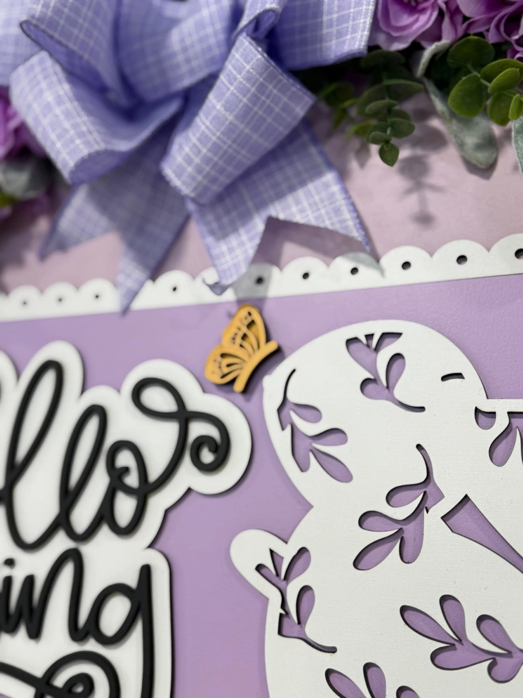Close-up of a lavender and white decorative sign with floral cutouts and the words Hello Spring in cursive. A wooden butterfly decoration is attached above. The sign is adorned with a purple gingham bow and greenery, creating a cheerful, seasonal ambiance.