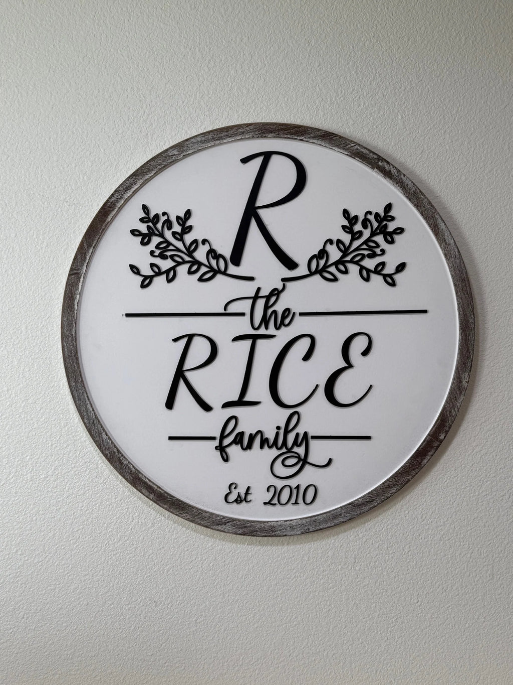 The Personalized Family Name Sign, Monogram Sign features a gray-trimmed round design with a white center, ornate black R, floral accents, the RICE family in mixed fonts, and “Est 2010”—a perfect custom decor piece.