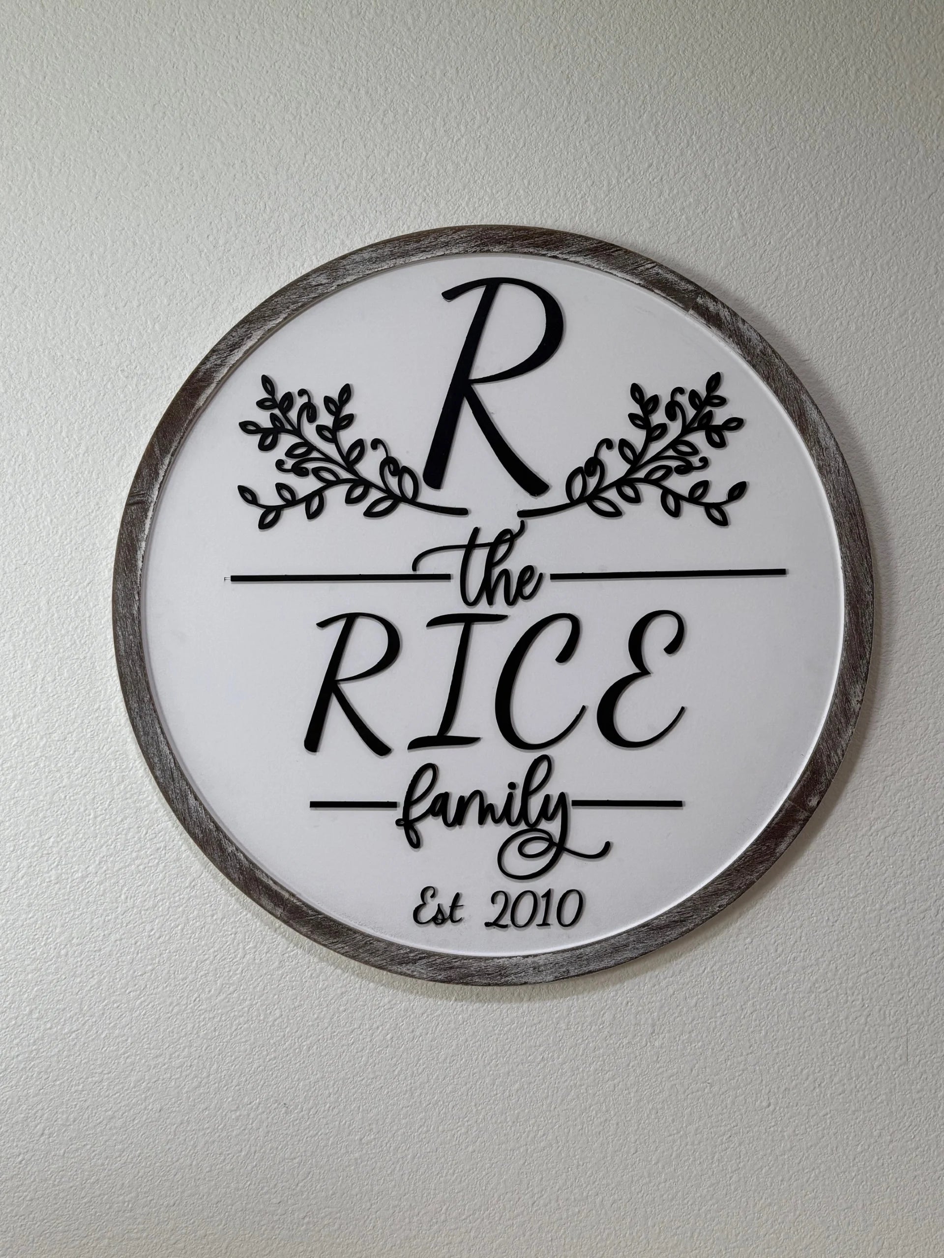 The Personalized Family Name Sign, Monogram Sign features a gray-trimmed round design with a white center, ornate black R, floral accents, the RICE family in mixed fonts, and “Est 2010”—a perfect custom decor piece.