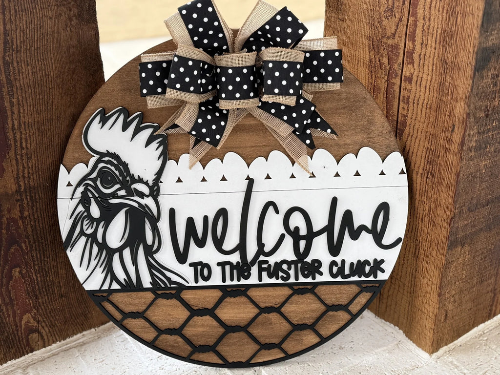 Round wooden sign with a hen illustration and text, Welcome to the Fuster Cluck. It features a decorative black and white polka dot bow with beige trim at the top. The bottom half is designed with a faux black wire fence pattern. The background is a wood surface.