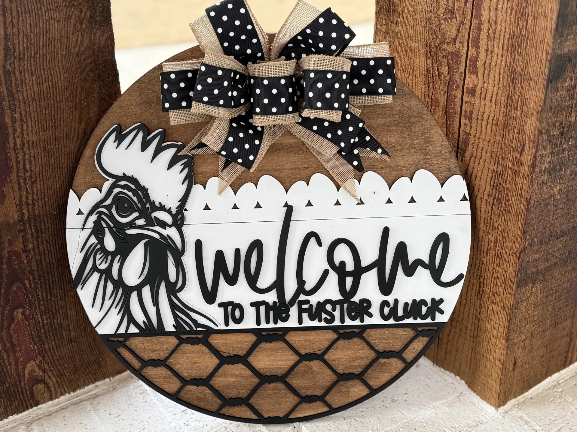 Round wooden sign with a hen illustration and text, Welcome to the Fuster Cluck. It features a decorative black and white polka dot bow with beige trim at the top. The bottom half is designed with a faux black wire fence pattern. The background is a wood surface.
