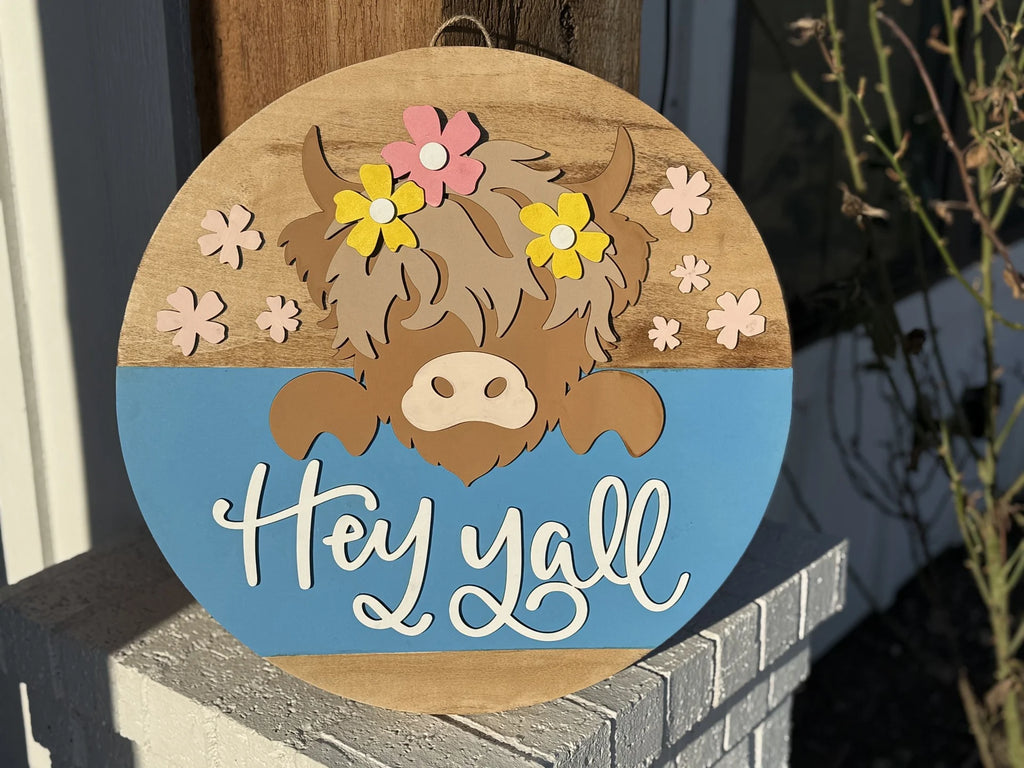 Round wooden sign with a cartoon cow face wearing pink and yellow flowers on its head. The top half is wood-toned with pink flowers, and the bottom half is blue with “Hey y’all” in white script. It hangs against a brick wall with plants in the background.