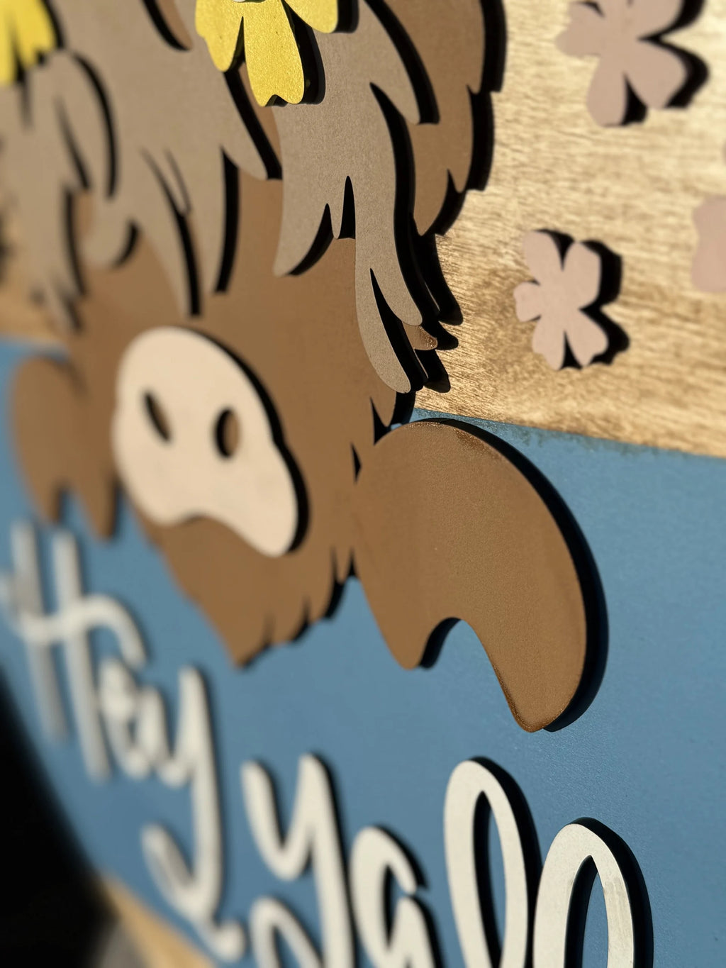 Close-up of a decorative sign featuring a stylized brown highland cow with two yellow flowers on its head and pink flowers around it. The background includes a smooth light brown top and a blue section at the bottom with the phrase Hey Yall in white cursive letters.