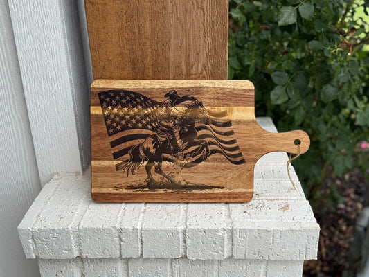 A wooden cutting board with an engraved image of a cowboy riding a bucking horse in front of a waving American flag, resting on a white brick surface with greenery in the background.