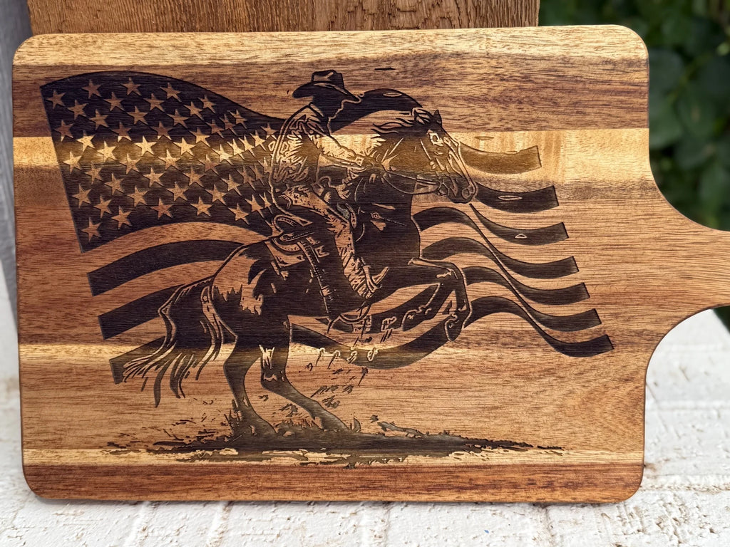 A wooden cutting board engraved with an image of a cowboy riding a rearing horse in front of a waving American flag.