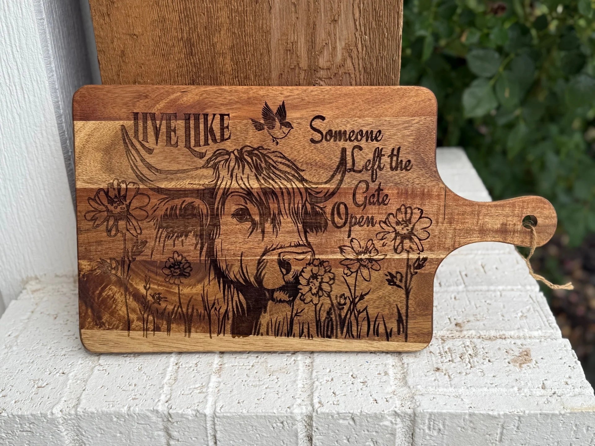 A wooden cutting board engraved with a cow, flowers, a bird, and the phrase “Live like someone left the gate open,” resting on a white brick ledge with greenery in the background.