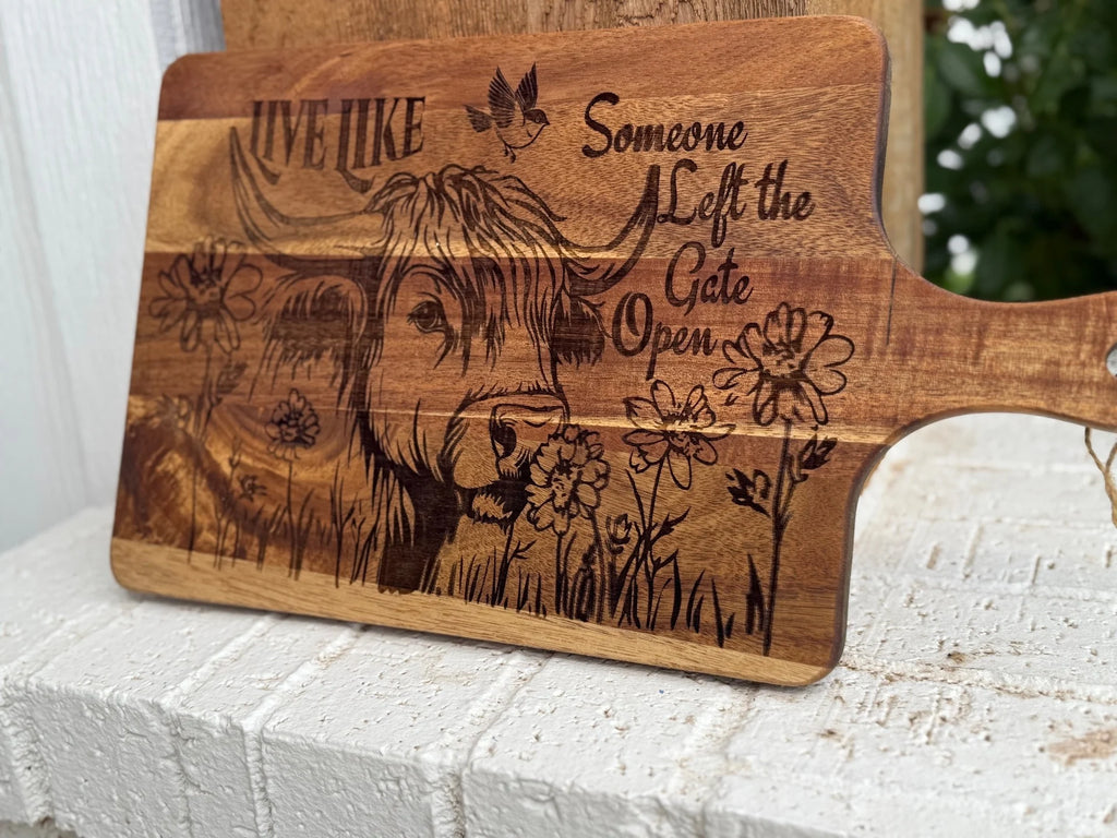 A wooden cutting board with an engraved illustration of a cow among flowers, a bird, and the text Live Like Someone Left the Gate Open. The board rests on a white brick surface.