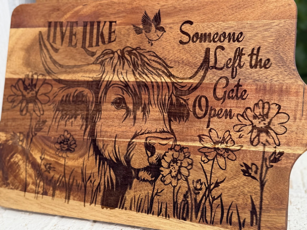A wooden board etched with an illustration of a cow among wildflowers, a bird above, and the words “Live like someone left the gate open” engraved in decorative script.