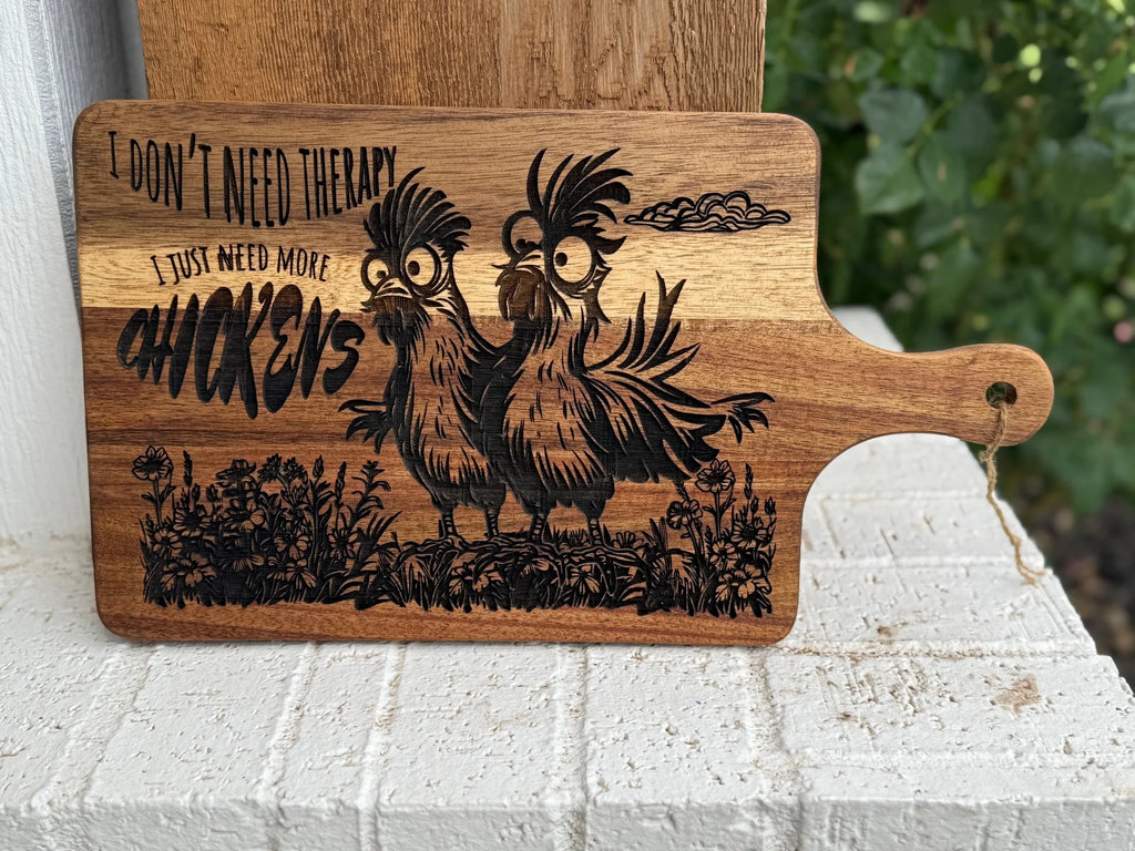 A wooden cutting board features a drawing of two quirky chickens and the text, I dont need therapy, I just need more chickens, engraved above and next to the birds. The board is outdoors, propped on a white brick surface.