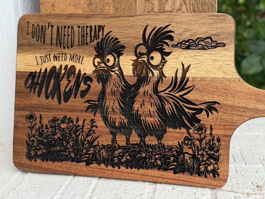 A wooden cutting board engraved with two quirky chickens and the text: I dont need therapy, I just need more chickens, surrounded by grass and flowers.