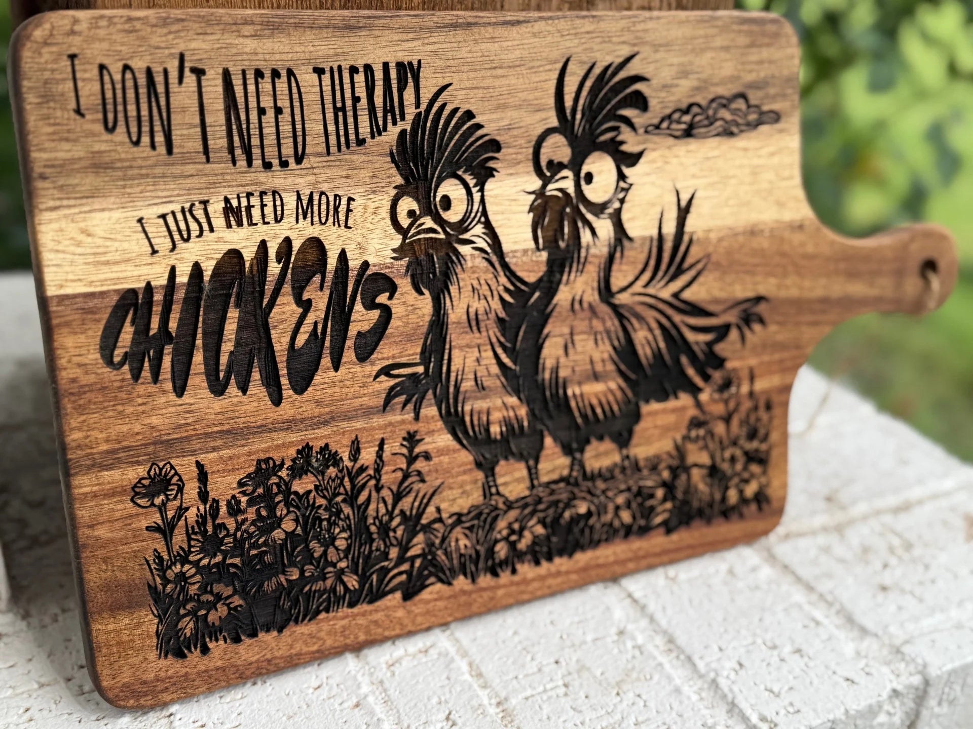 A wooden cutting board engraved with two quirky chickens standing in grass and flowers, alongside the text: I dont need therapy, I just need more chickens.
