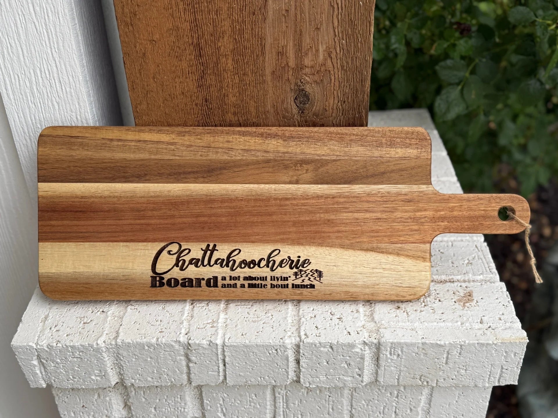 A wooden cutting board with the words Chattahoochee Board: a lot about livin and a little bout lunch engraved on it, rests on a white brick ledge with greenery in the background.