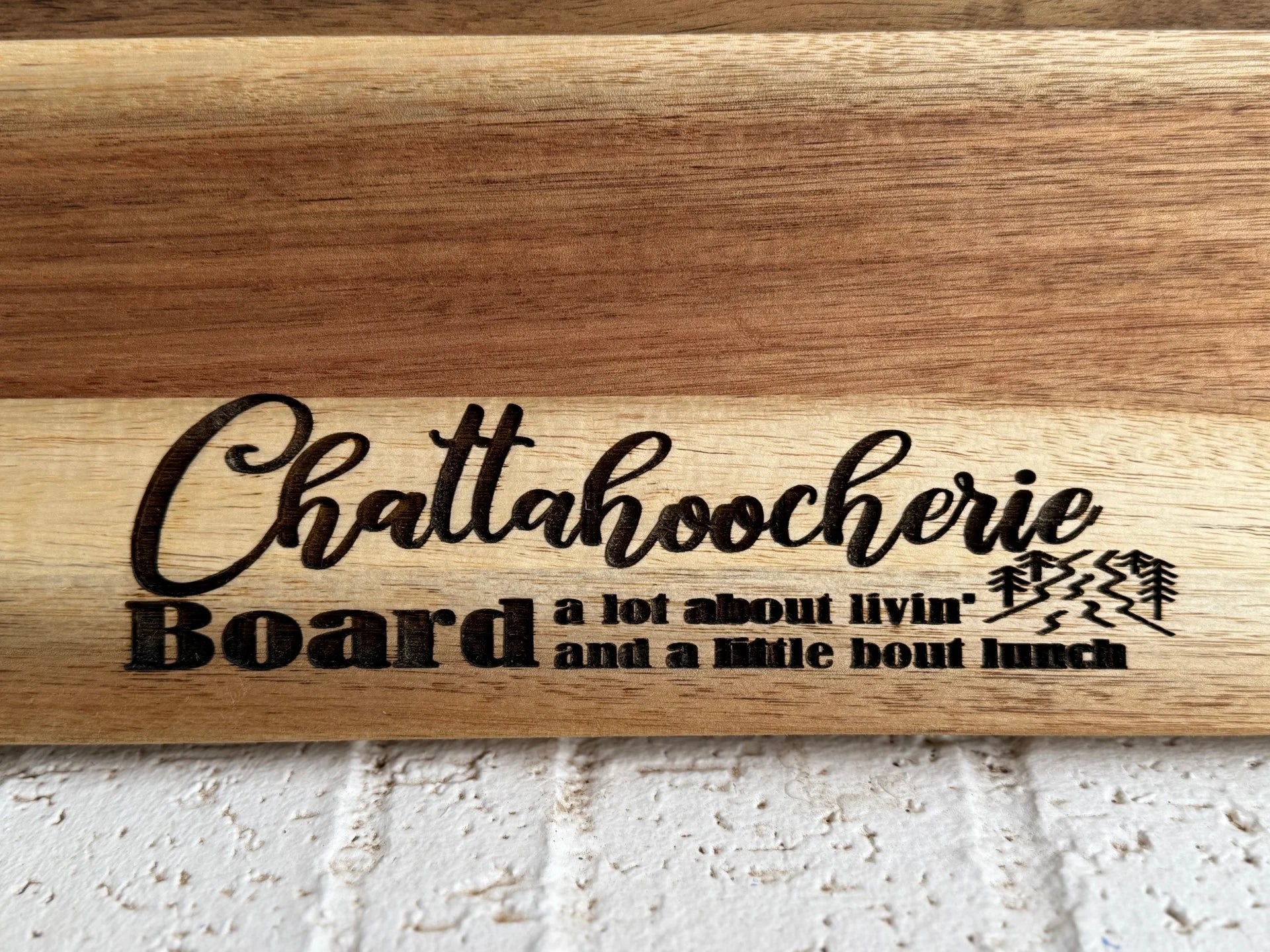 A wooden board engraved with the words Chattahoochee Board a lot about livin and a little bout lunch, along with a small image of two trees and a river.