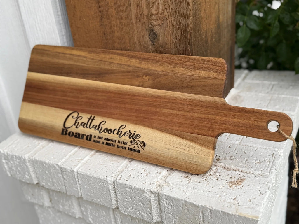 A wooden serving board with the words Chattahoocheerie Board: a lot about livin’ and a little bout lunch engraved on it, resting on a white brick ledge outdoors near some greenery.