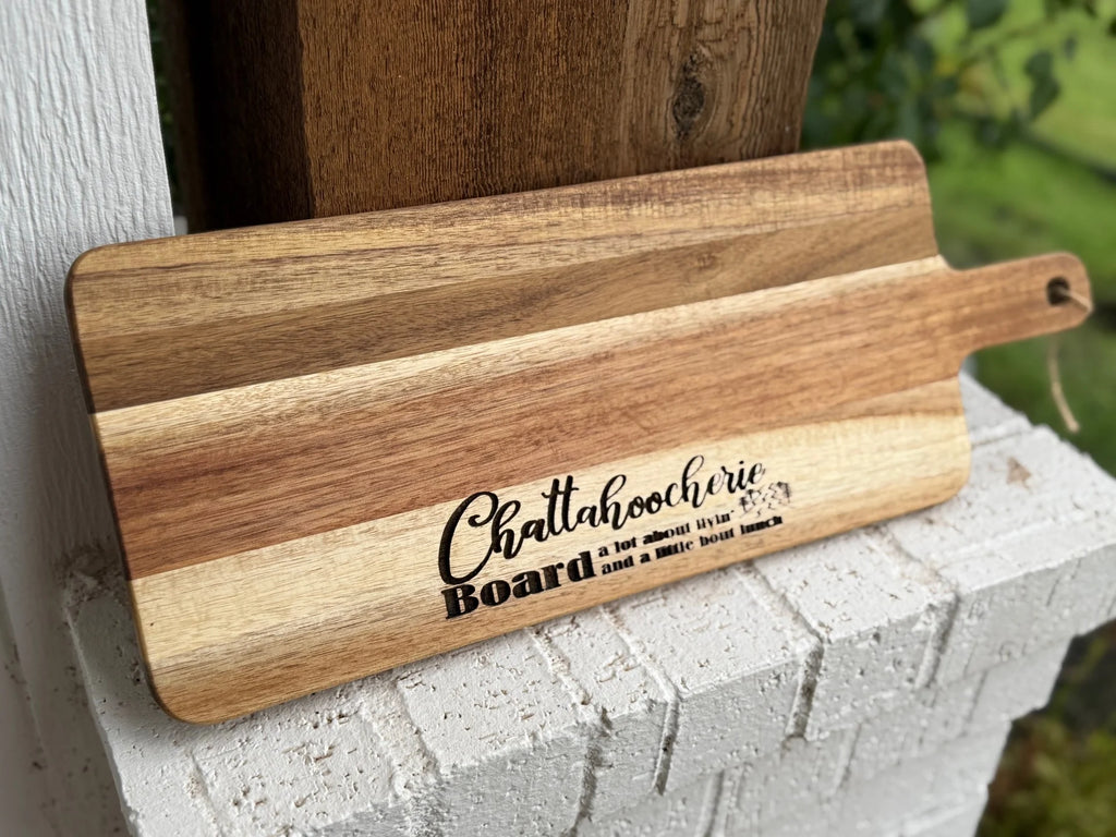 A wooden charcuterie board with the words Chattahoocheerie Board and a playful phrase printed on it, resting on a white brick surface outdoors.