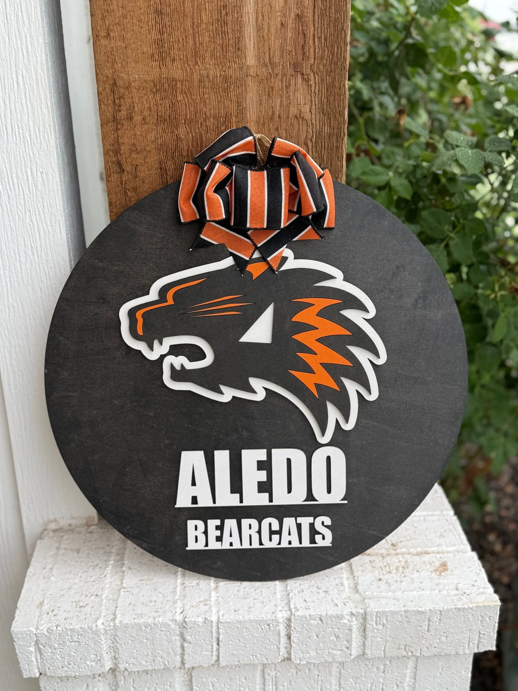 The Aledo Bearcat Mascot Door Hanger Door Sign is a round black sign with the orange and white Aledo Bearcat logo, Aledo Bearcats text, and an orange-black striped ribbon, perfect for displaying school spirit outdoors.