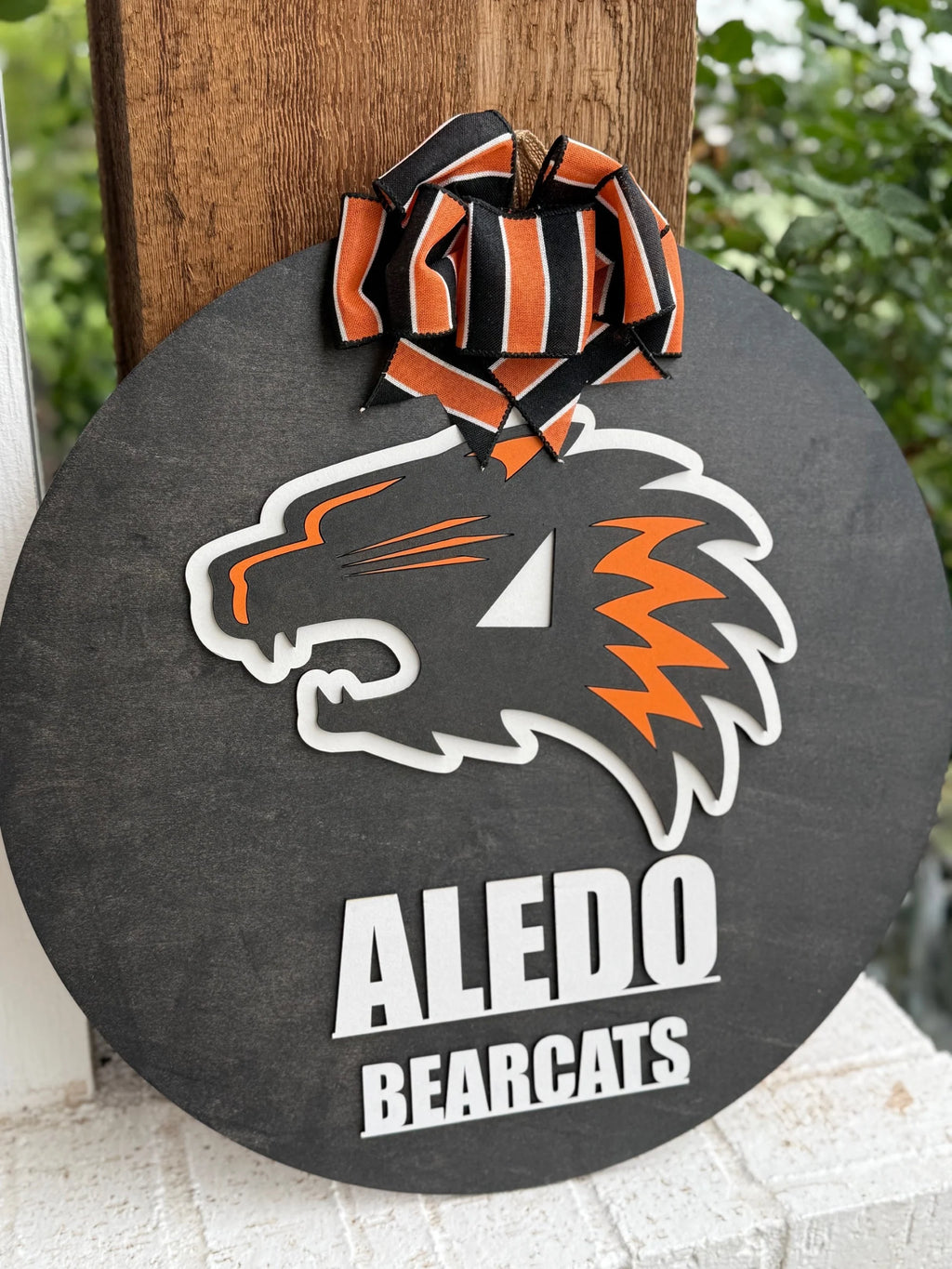 The Aledo Bearcat Mascot Door Hanger Door Sign is a round black door sign with an orange and white Aledo Bearcat logo, bold Aledo Bearcats text, and a striped ribbon bow—perfect for any fan’s door.