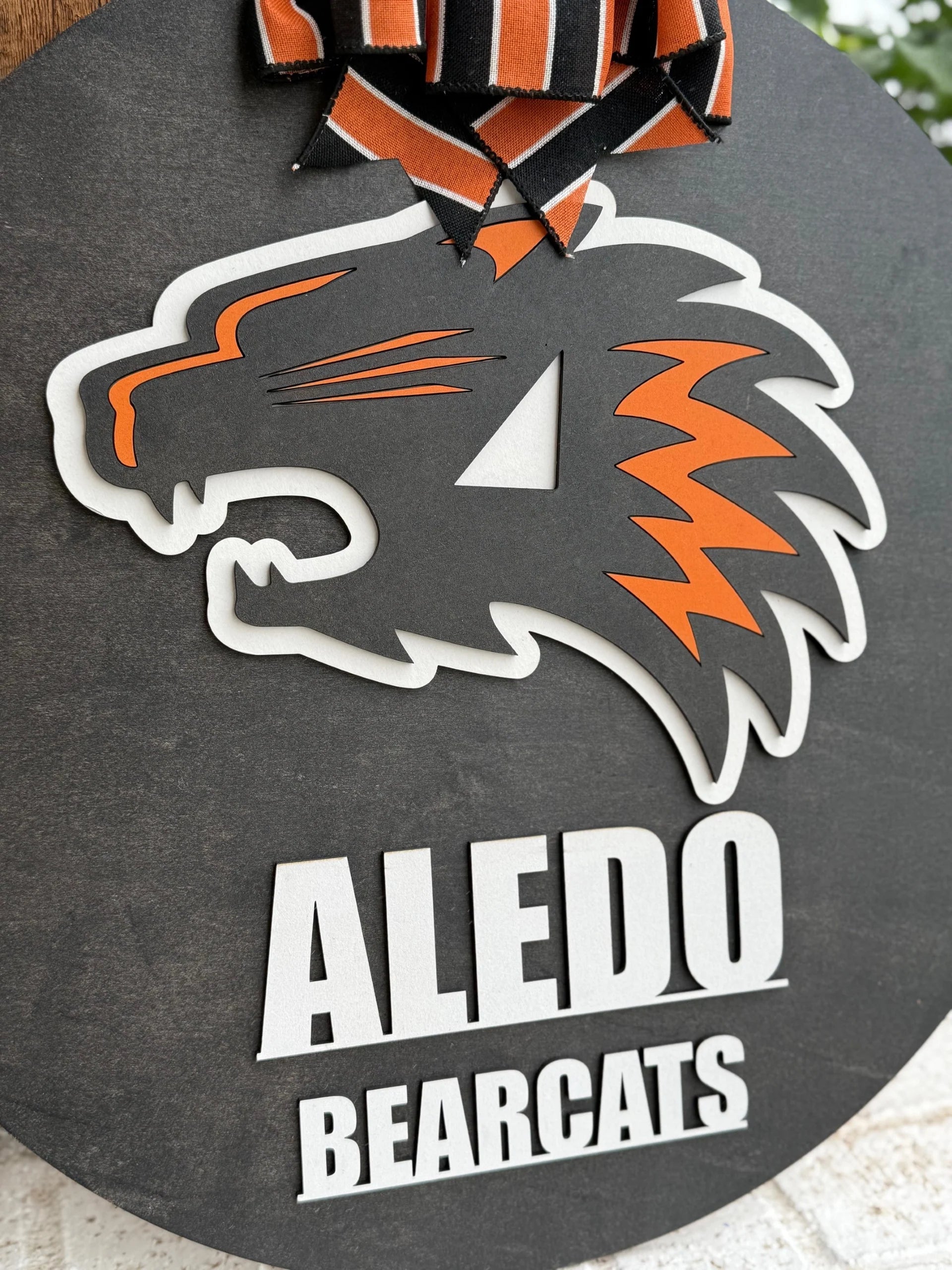 The Aledo Bearcat Mascot Door Hanger Door Sign is a round black sign featuring a bold orange, white, and black bearcat logo with Aledo Bearcats below. Finished with a black and orange striped bow, it’s perfect for showcasing team spirit on your door.
