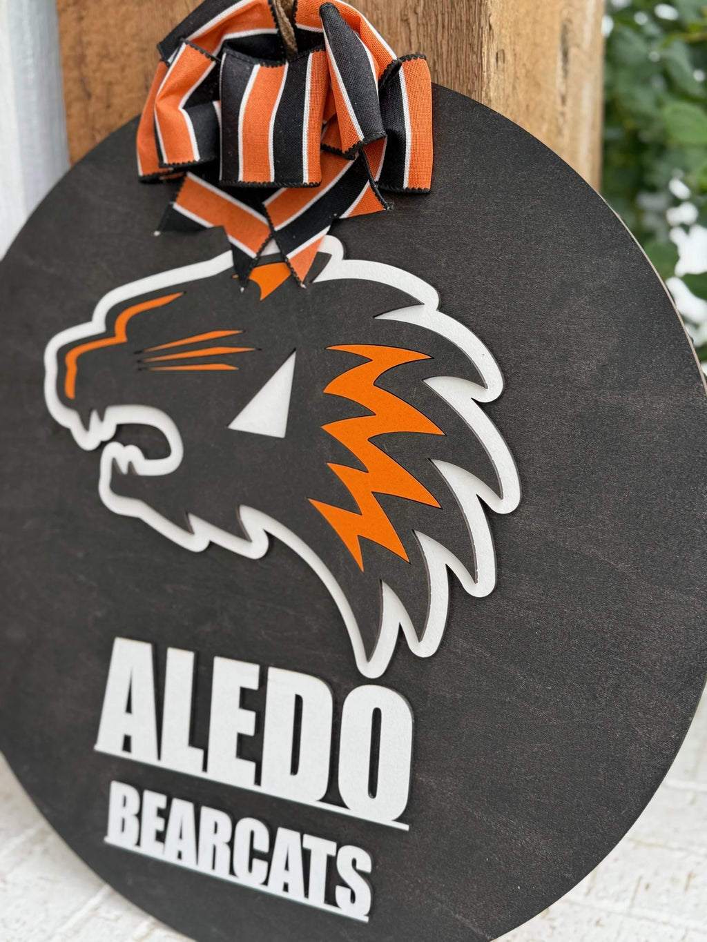 The Aledo Bearcat Mascot Door Hanger Door Sign is a round black hanger with an orange and white bearcat logo, Aledo Bearcats text, and a striped ribbon in black, orange, and white, shown hanging on wood.