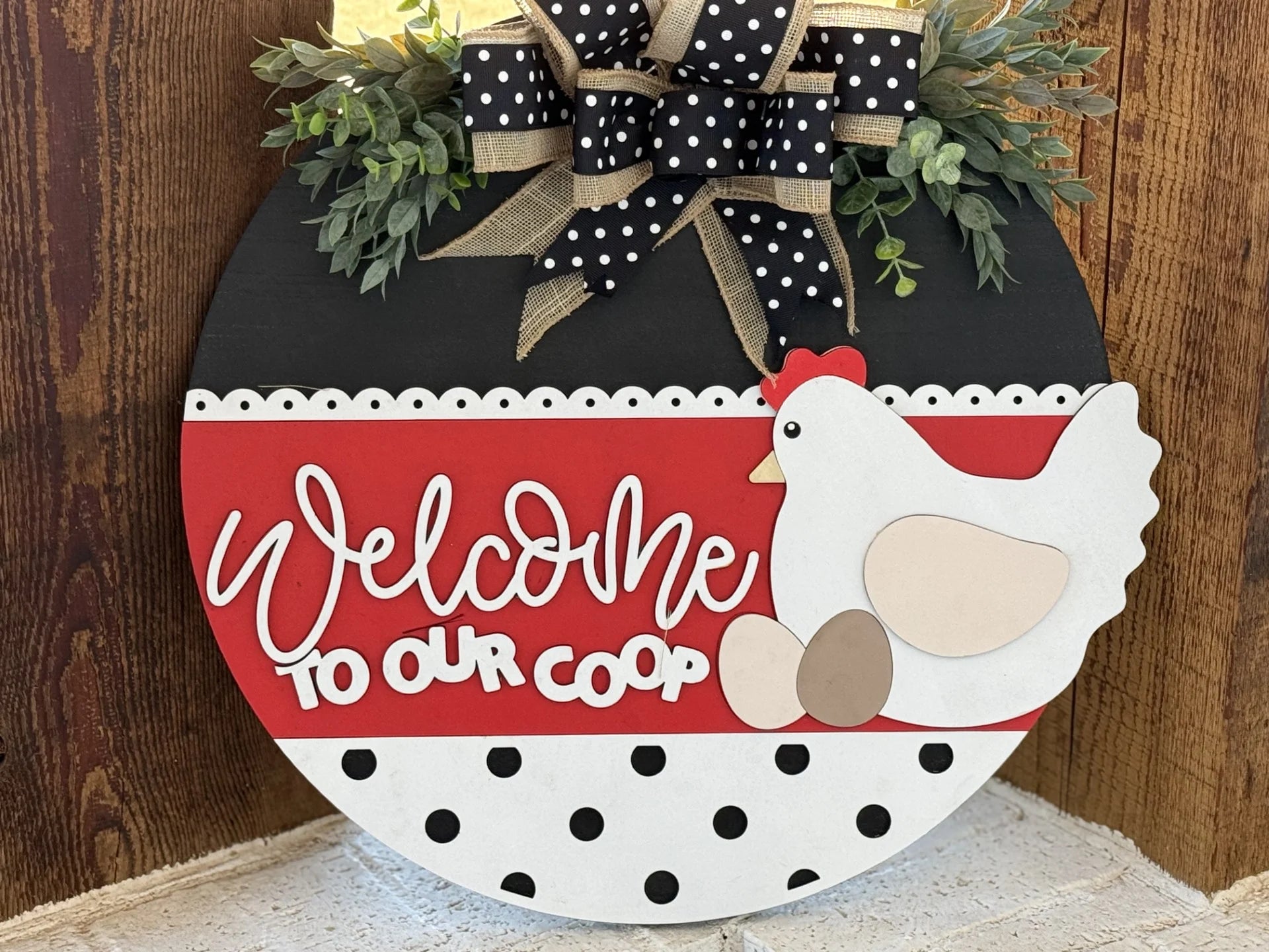 A circular sign named If Were Not Home Check the Rink Door Sign features a chicken and eggs design with Welcome to Our Coop in white script on red. It acts as a charming wreath with a black top, greenery, and polka dot bow, leaning against a wood-paneled wall.