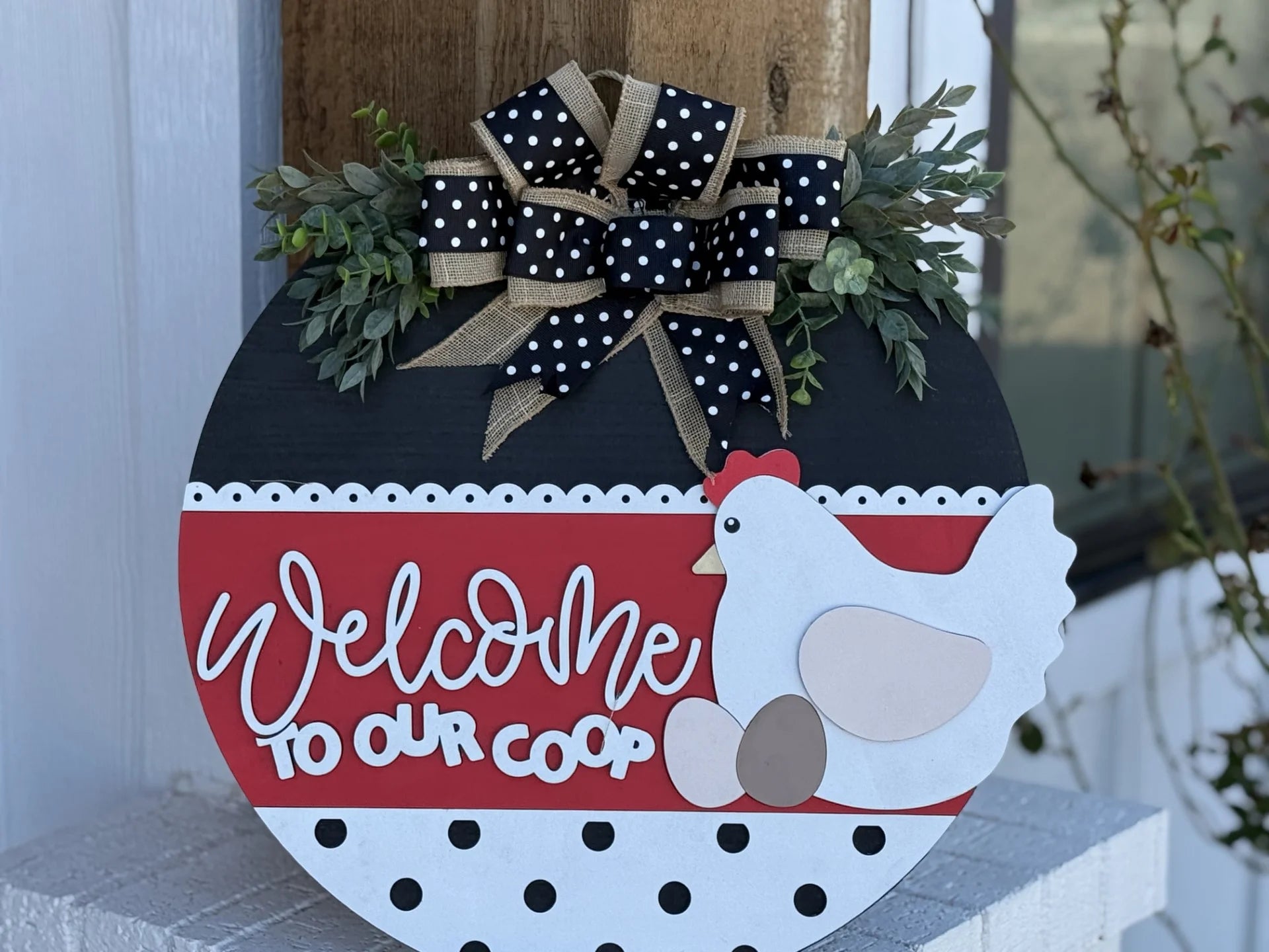 The If Were Not Home Check the Rink Door Sign features a white chicken and eggs on a red and black background. It dons a polka dot burlap bow and greenery, serving as charming home decor on an exterior wooden wall near a window—ideal for farmhouse enthusiasts.