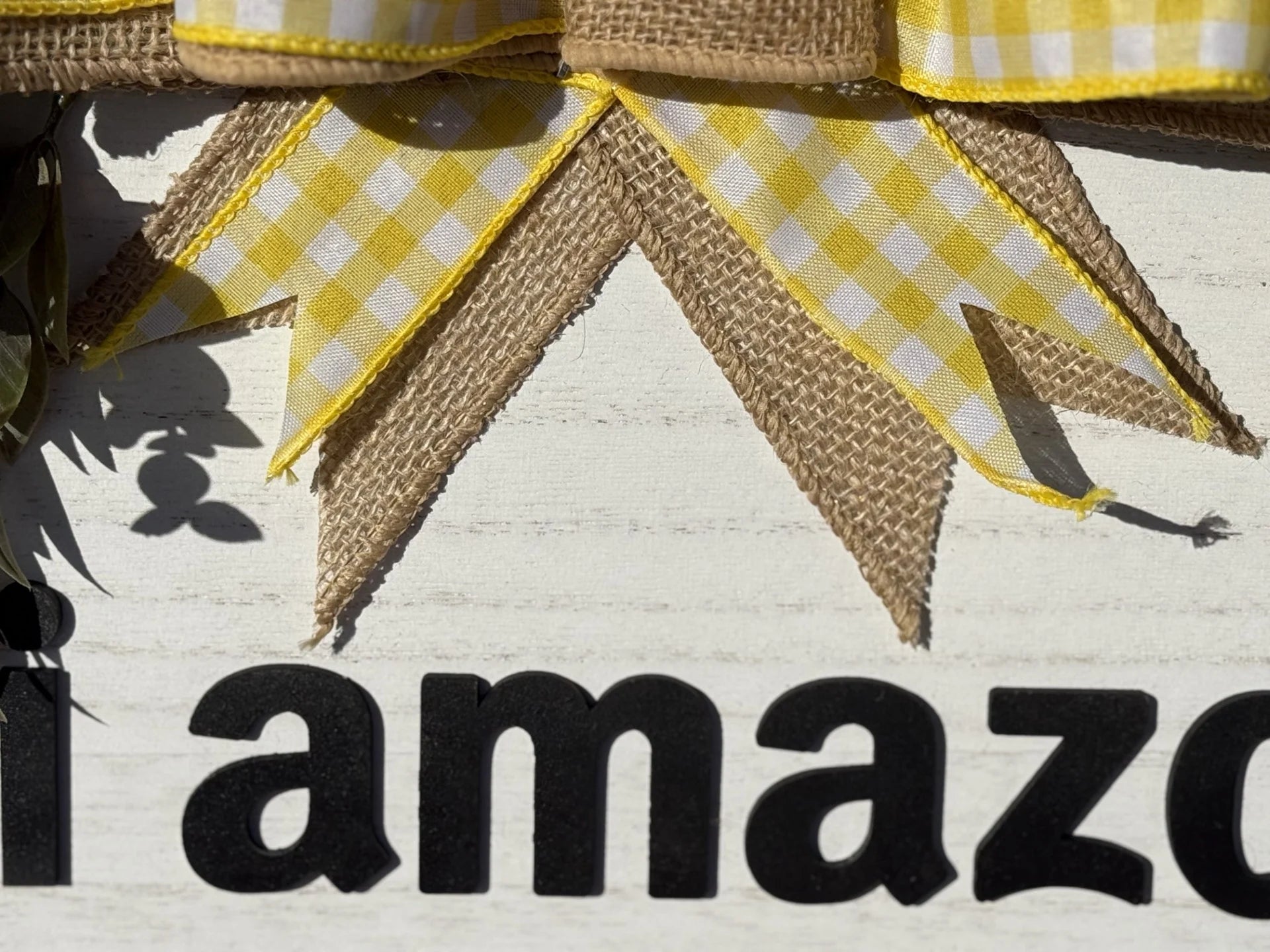 A close-up of the Hi Amazon Yes This House Again door sign against a white wooden background features a burlap and yellow gingham ribbon at the top, framing partial black text i amazo. Sunlight and shadows enhance this Amazon-themed creation.