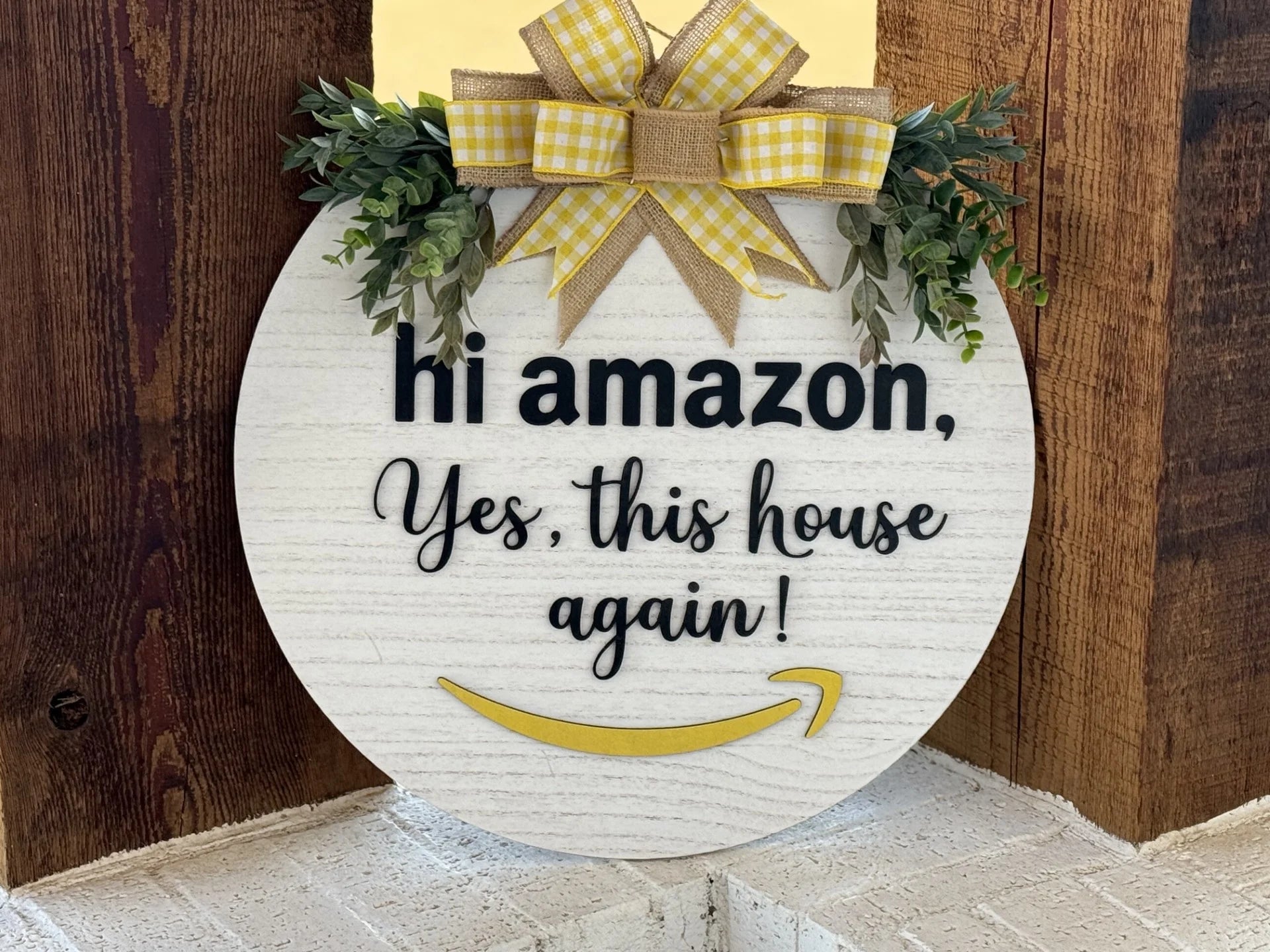The Hi Amazon Yes This House Again Door Sign is a unique wooden wreath with a whitewashed finish, featuring the phrase in black script. Its accented with a decorative bow of yellow plaid, burlap ribbons, green foliage, and completed by a playful yellow smiley-shaped arrow.