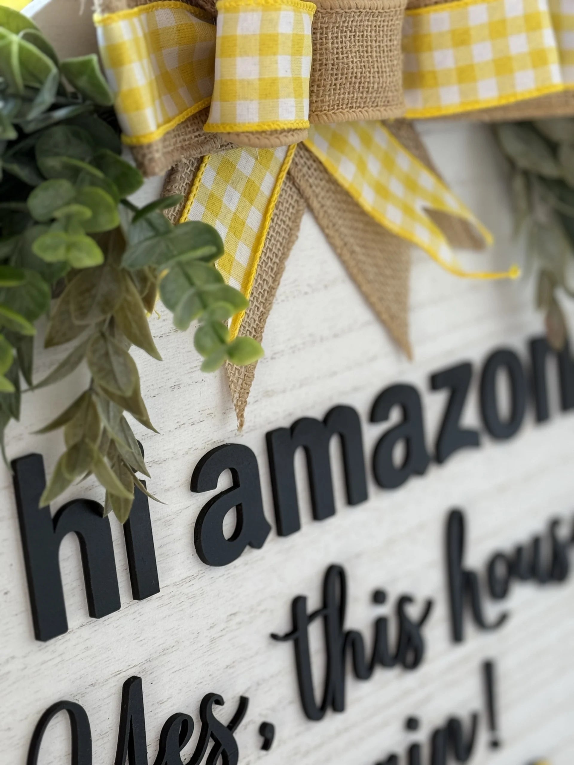 Close-up of the Hi Amazon Yes This House Again Door Sign on a white background with black text, adorned with a burlap and yellow gingham ribbon bow and greenery accents. The text reads hi Amazon, and Yes, this hous, with part of the message partially obscured.