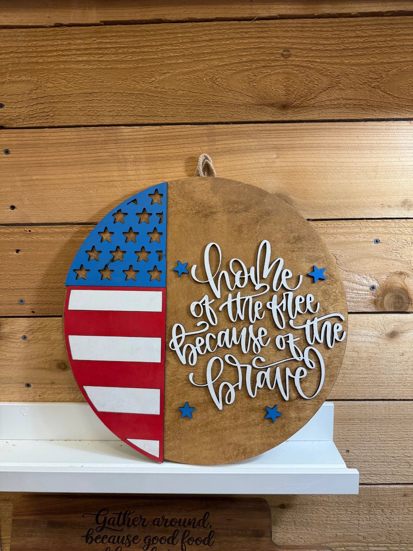 A round wooden sign with a partial American flag on the left and script text on the right reading Home of the free because of the brave, hanging on a wood wall above a white shelf.