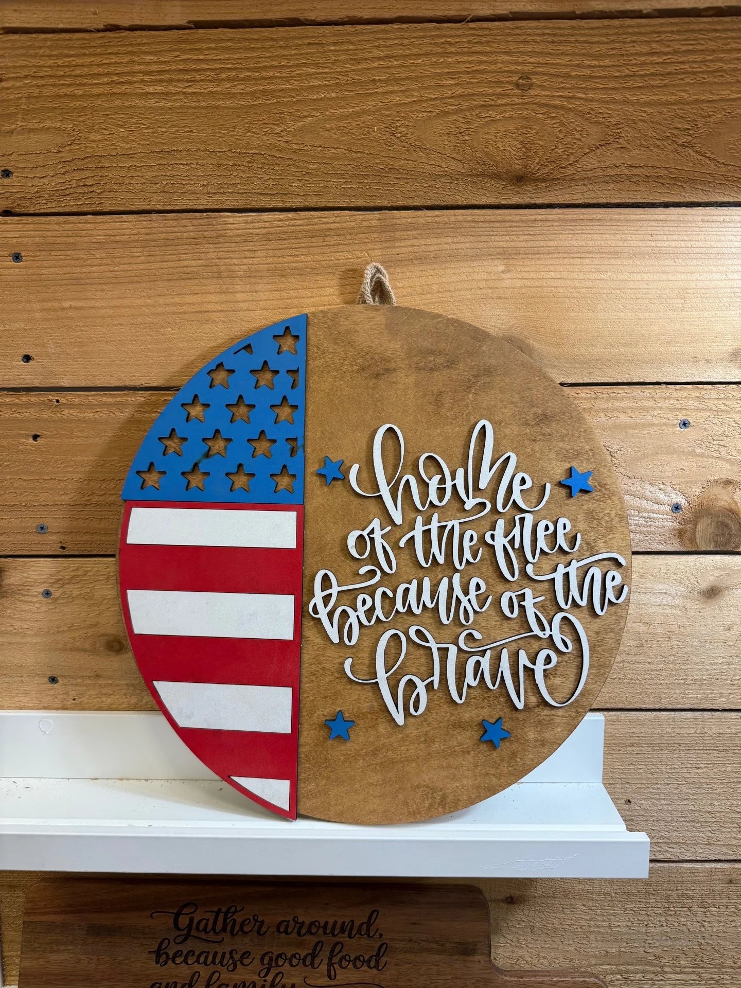 A round wooden sign with a U.S. flag design on the left and the words Home of the free because of the brave in white script on the right, hanging on a wooden wall.