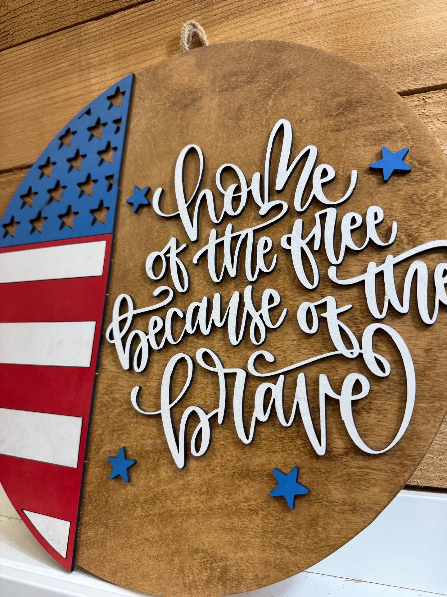 A wooden wall hanging features a partial American flag on the left and white script text reading Home of the free because of the brave with blue stars on a round wood background.