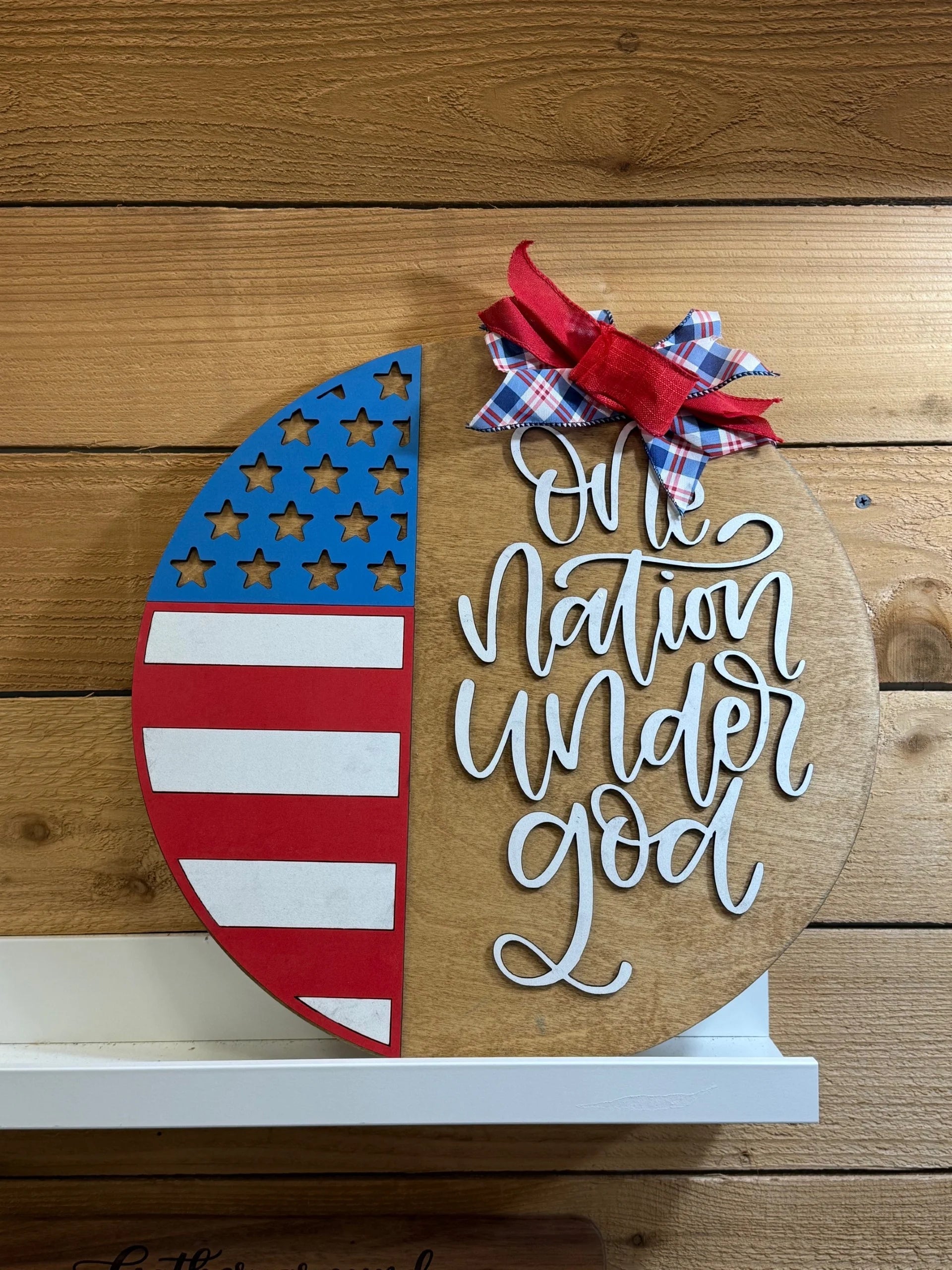 A wooden sign with half painted as the American flag and the other half reading One nation under God in white script, topped with a red, white, and blue bow, mounted on a wooden wall.