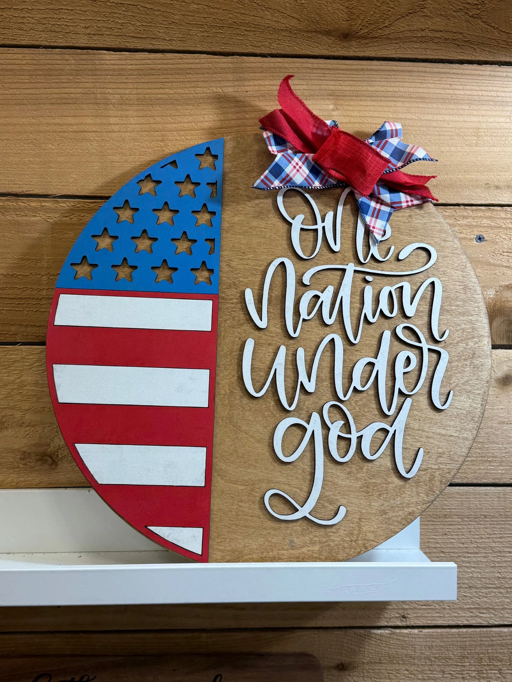 A round wooden sign with half painted as the American flag and the other half reading One nation under God in white script. A red and plaid bow is attached at the top right. The sign is against a wood wall.
