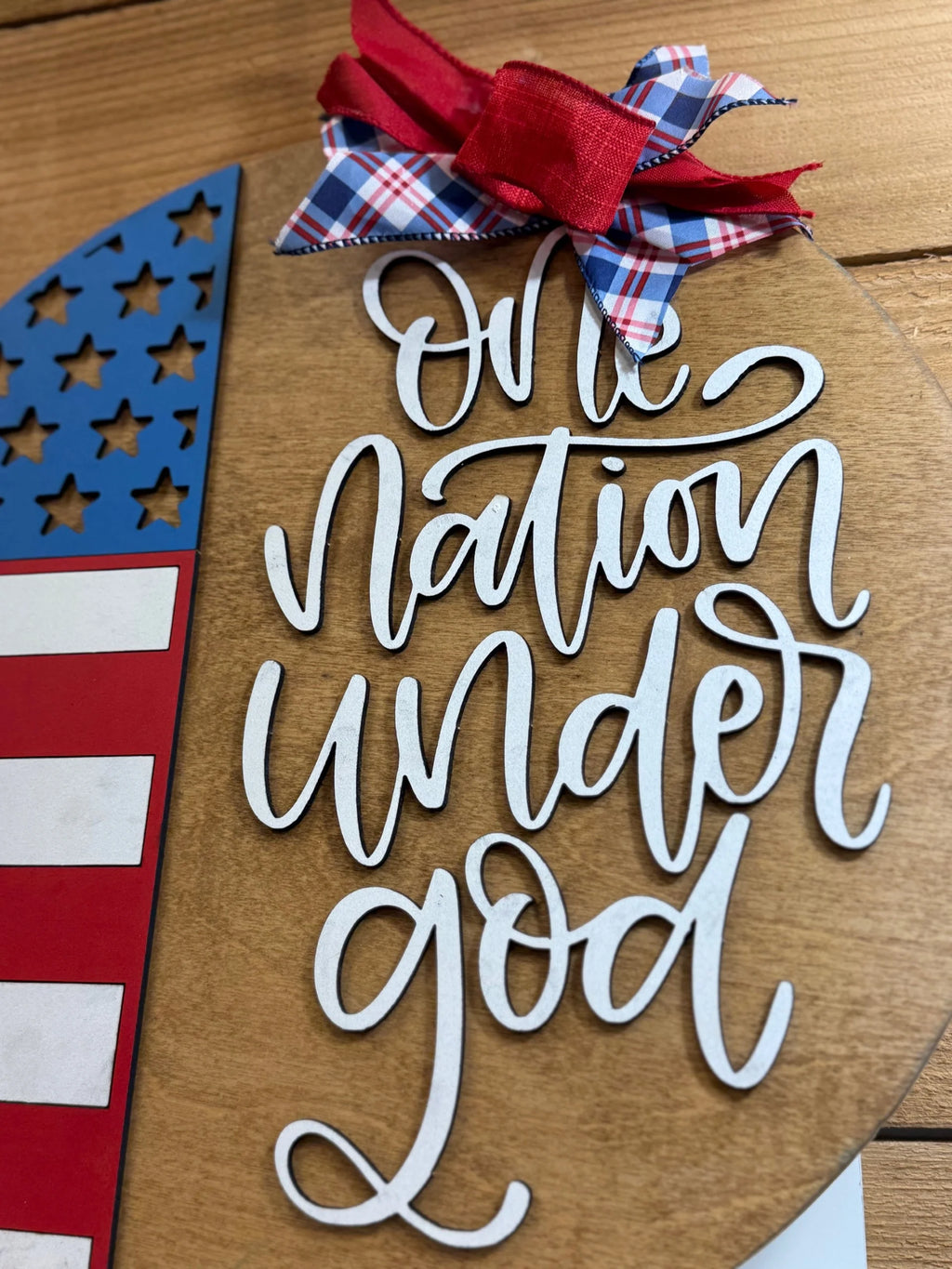 A wooden sign features a partial American flag and the words One Nation Under God in white script, decorated with a red and plaid bow, set against a wood background.