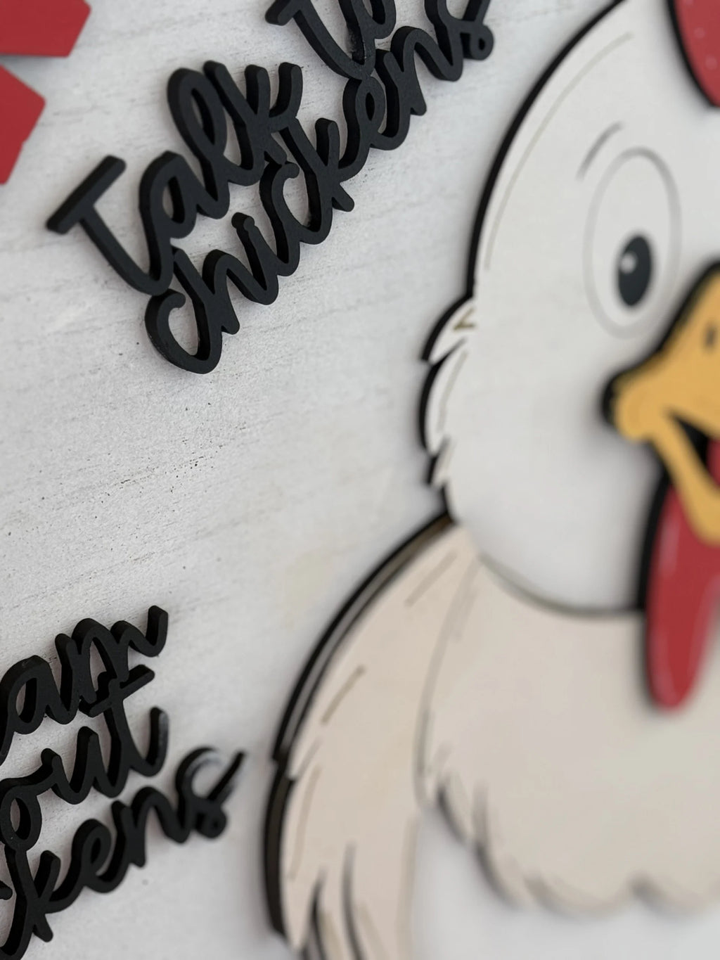 Close-up of a wall decoration featuring a cartoon chicken with a white body and red comb, looking to the side. Raised black text reads Talk to chickens above and learn about chickens below. The background is a textured, light gray surface.