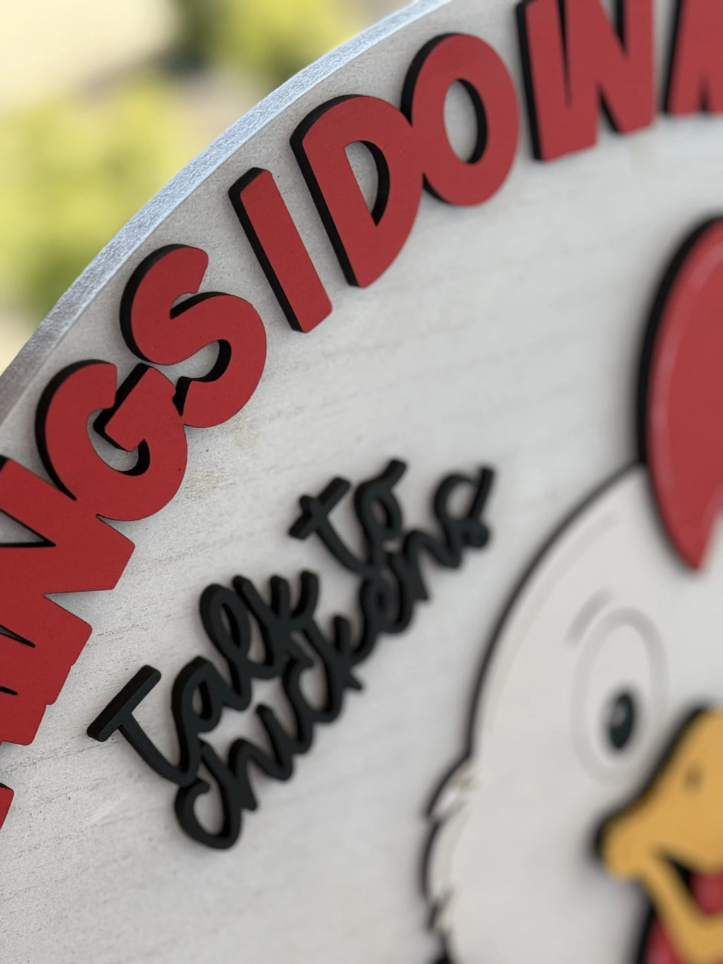 Close-up of a colorful sign featuring a cartoon chicken with a red comb and yellow beak. The prominent red text curves along the top edge, partially reading THINGS I DO W, while black text below reads Talk to chickens in a playful font. Background is blurred greenery.