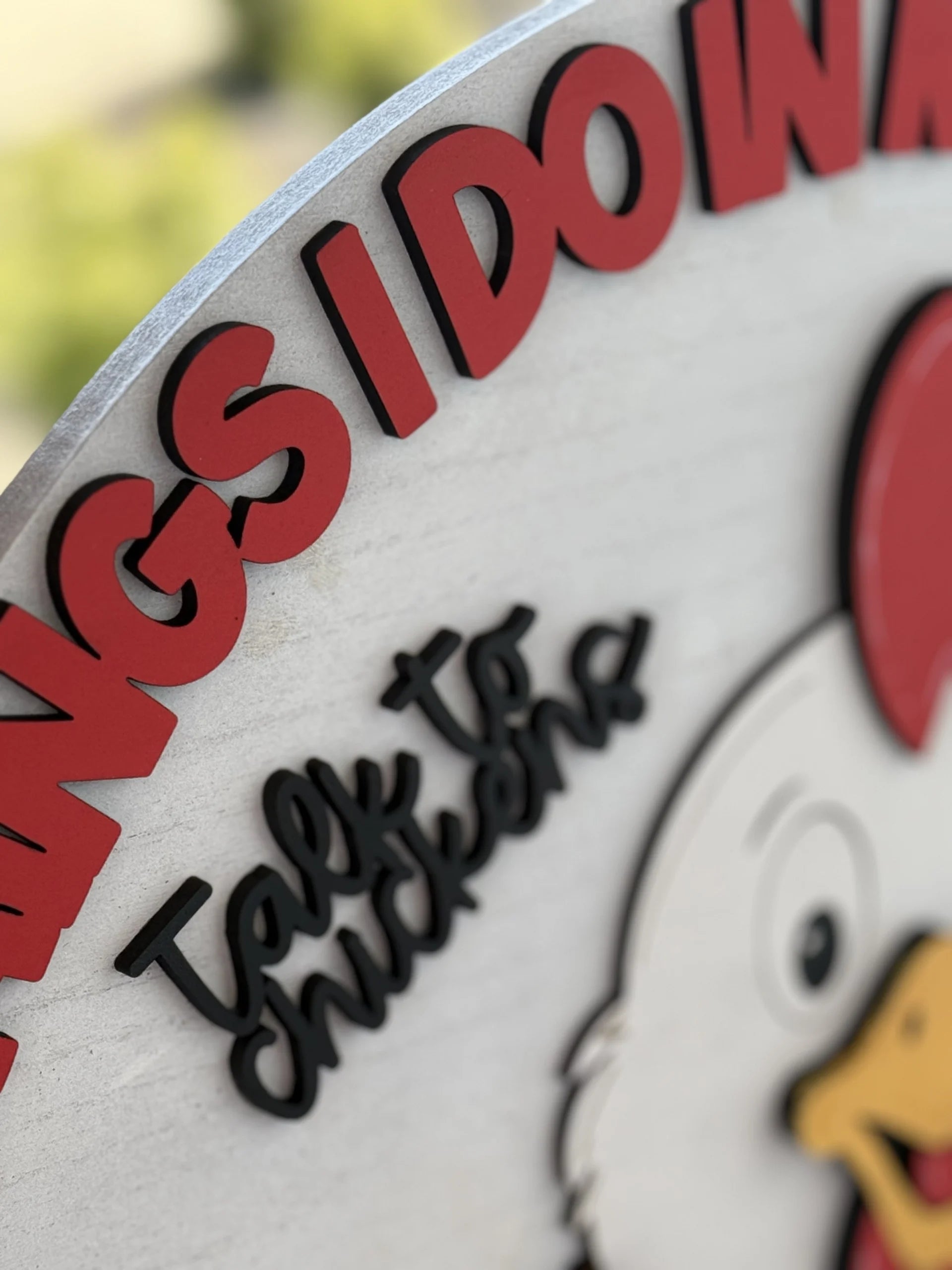 Close-up of a colorful sign featuring a cartoon chicken with a red comb and yellow beak. The prominent red text curves along the top edge, partially reading THINGS I DO W, while black text below reads Talk to chickens in a playful font. Background is blurred greenery.