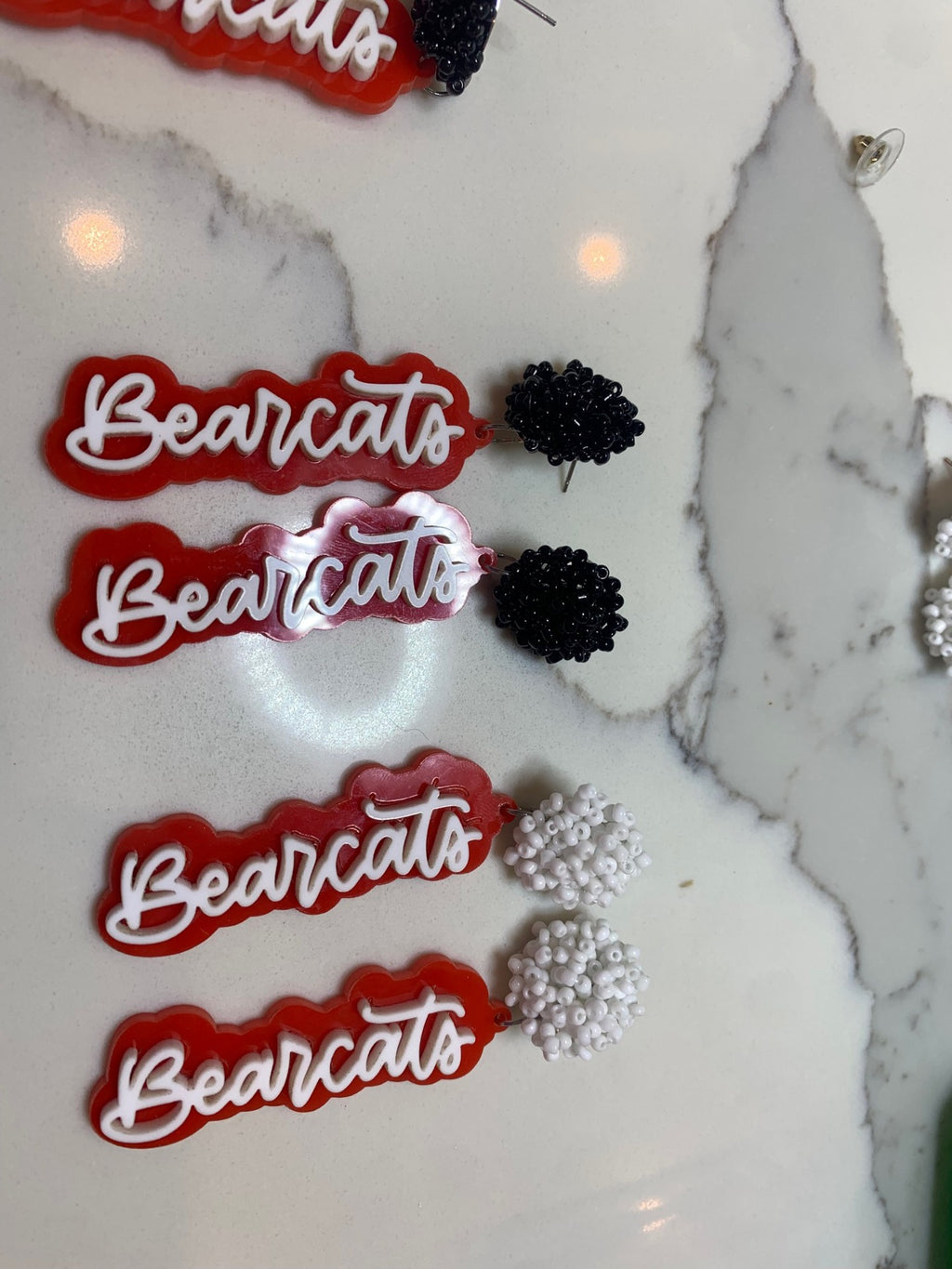 Set on a marble surface, the Aledo Bearcat Earrings showcase four red and white elements with Bearcats script beside black and white flower-shaped beaded clusters. The shiny, textured design of the clusters effortlessly captures attention.