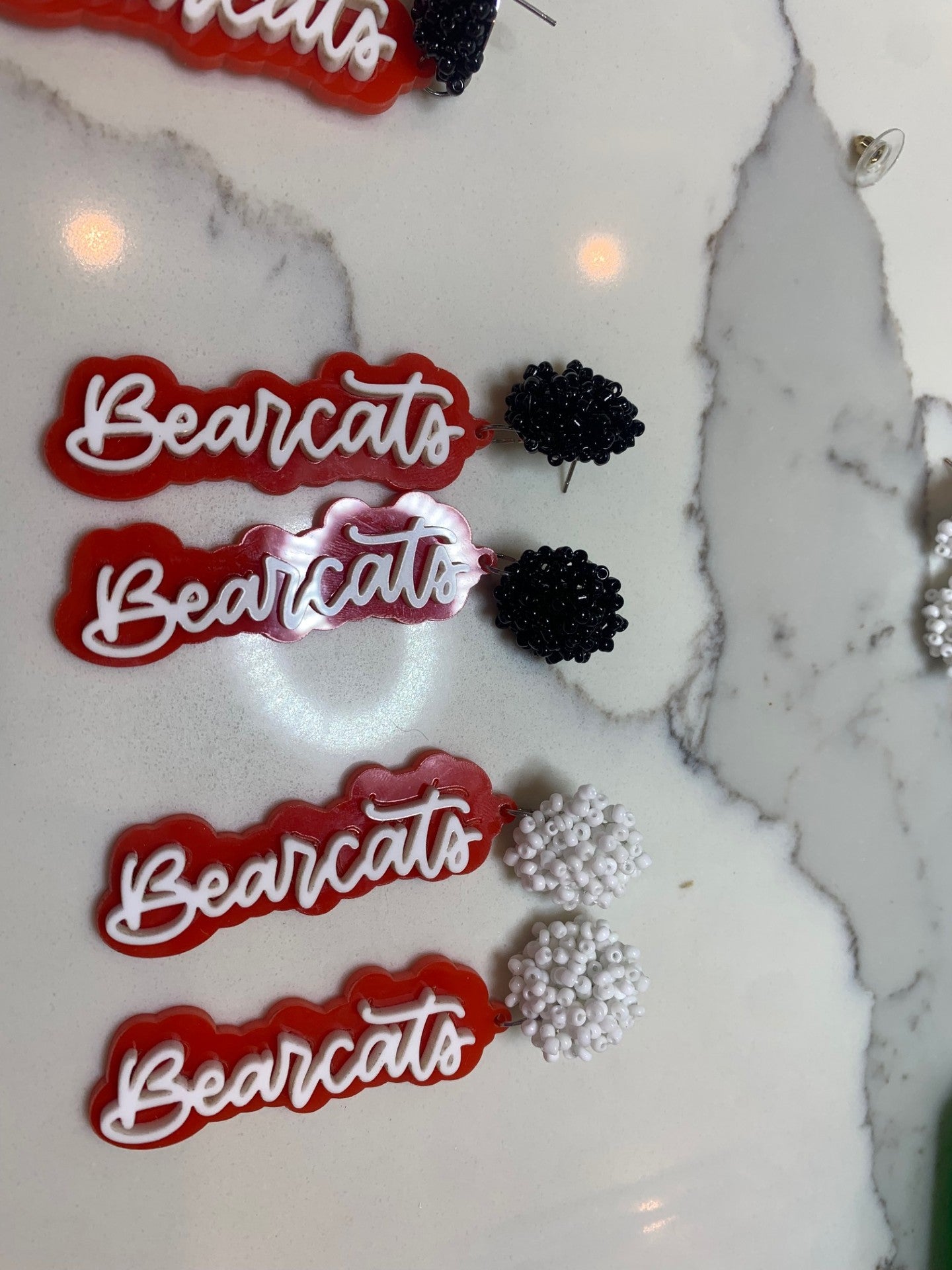 Set on a marble surface, the Aledo Bearcat Earrings showcase four red and white elements with Bearcats script beside black and white flower-shaped beaded clusters. The shiny, textured design of the clusters effortlessly captures attention.