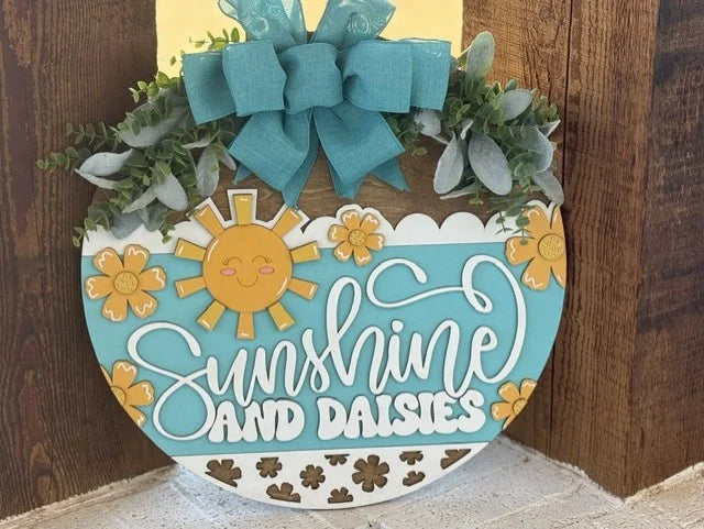 A decorative round sign with a cheerful design. It features the text Sunshine and Daisies in white cursive on a blue background. Above, theres a cartoon sun and daisies. A teal bow and green foliage accent the top, with wooden texture visible behind the sign.