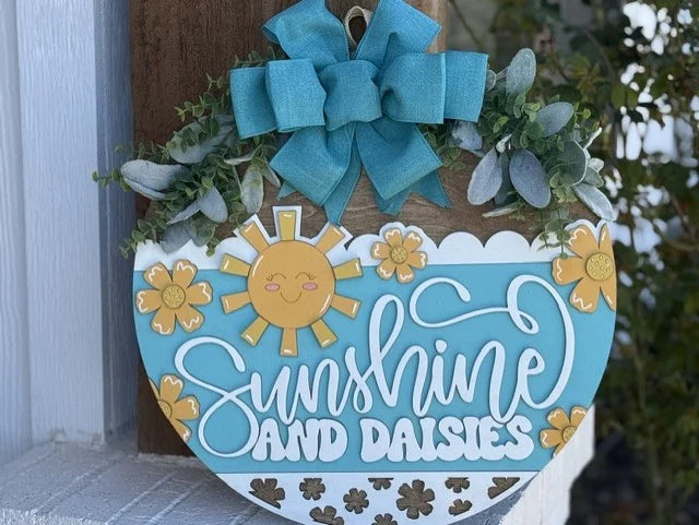 A heart-shaped wooden sign with the words Sunshine and Daisies in white cursive against a light blue background. It features a smiling sun and yellow daisies. A teal bow with greenery is attached on top. The sign is mounted on a wooden surface and rests near a white structure.