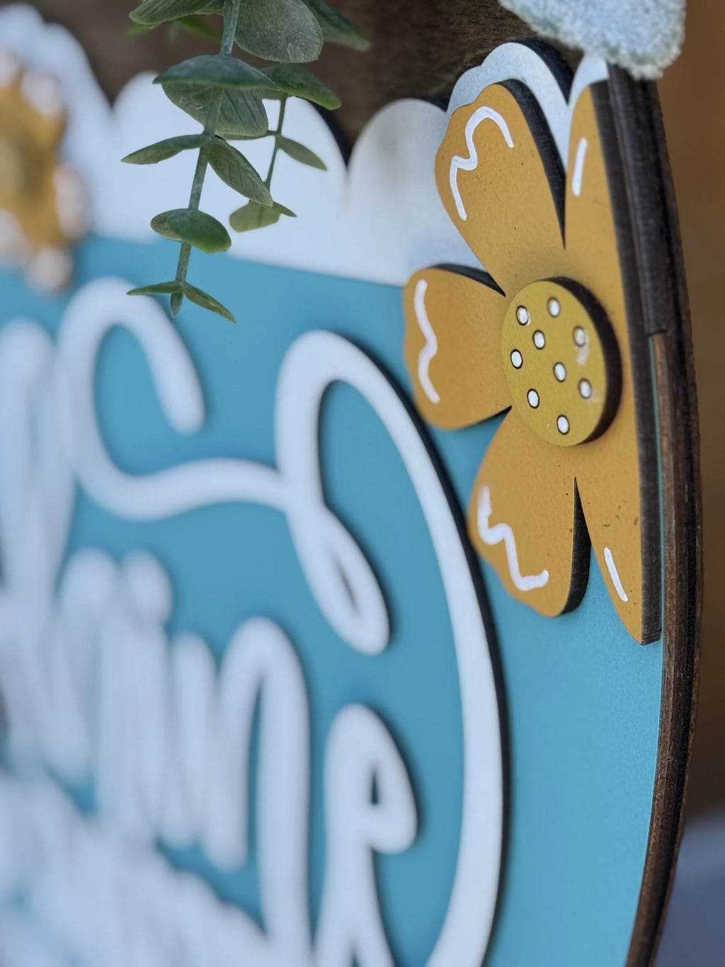 Close-up of a decorative sign featuring a yellow flower with white accents on a teal and white background. Green foliage extends from the top. The words on the sign are partially visible, displaying elegant, cursive lettering. The overall design has a cheerful and playful feel.