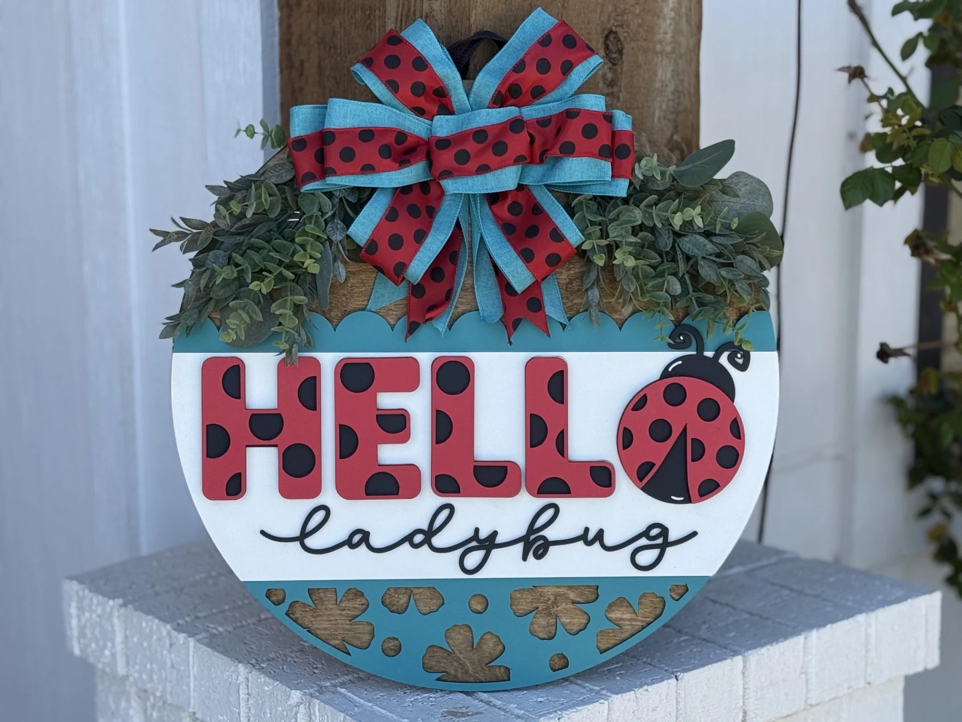 A circular wooden sign features Hello Ladybug in bold text, adorned with a ladybug motif. Its decorated with lush greenery and a large red bow with black polka dots. The background includes blue and white sections, and light brown clovers on the bottom. The sign is mounted on wood.