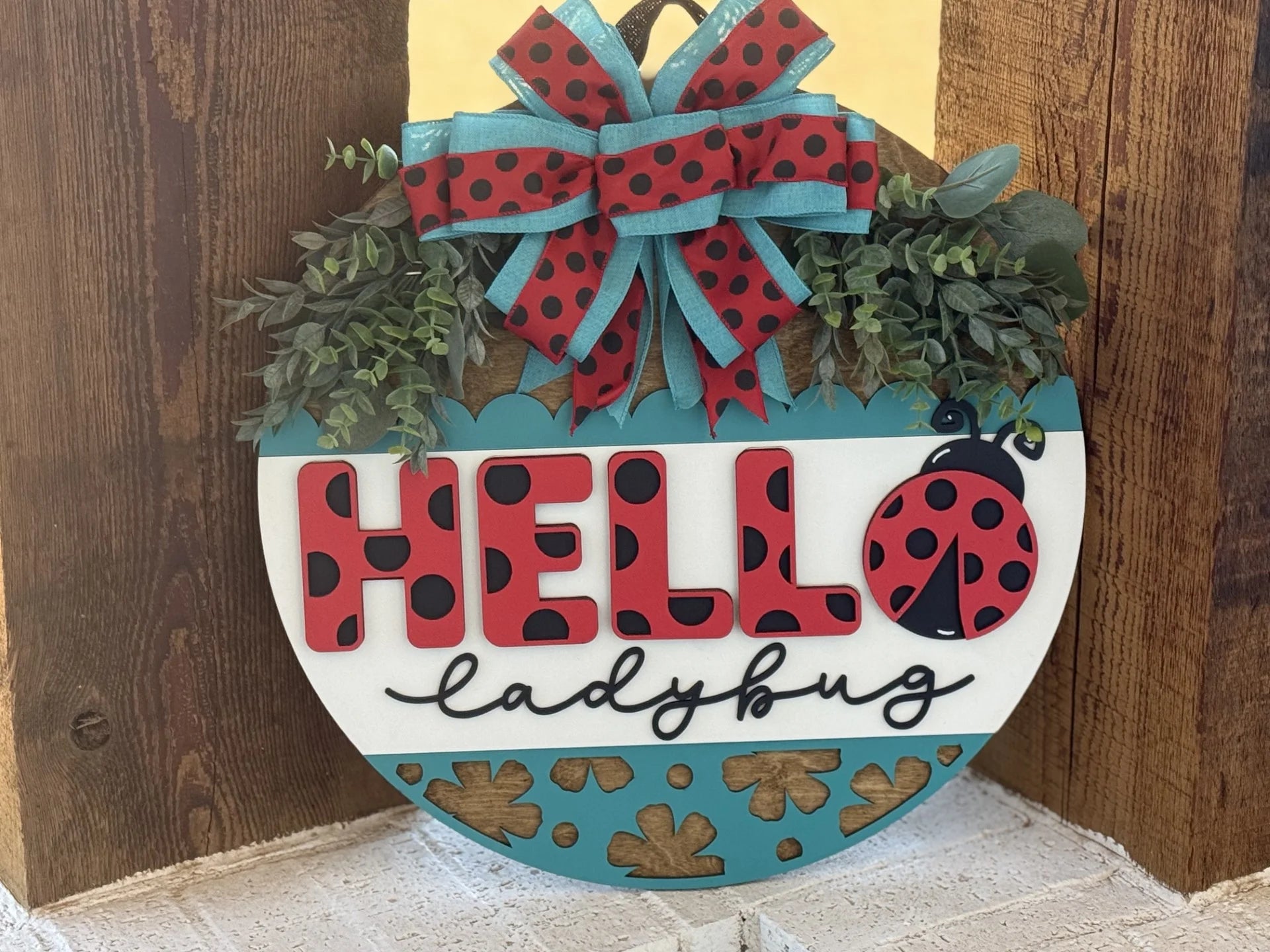 A decorative wooden sign with the text HELLO ladybug in red letters featuring ladybug spots. Above is a large bow with red and black spots and green ribbon. Greenery surrounds the bow, and cut-out leaf patterns adorn the bottom edge of the sign.