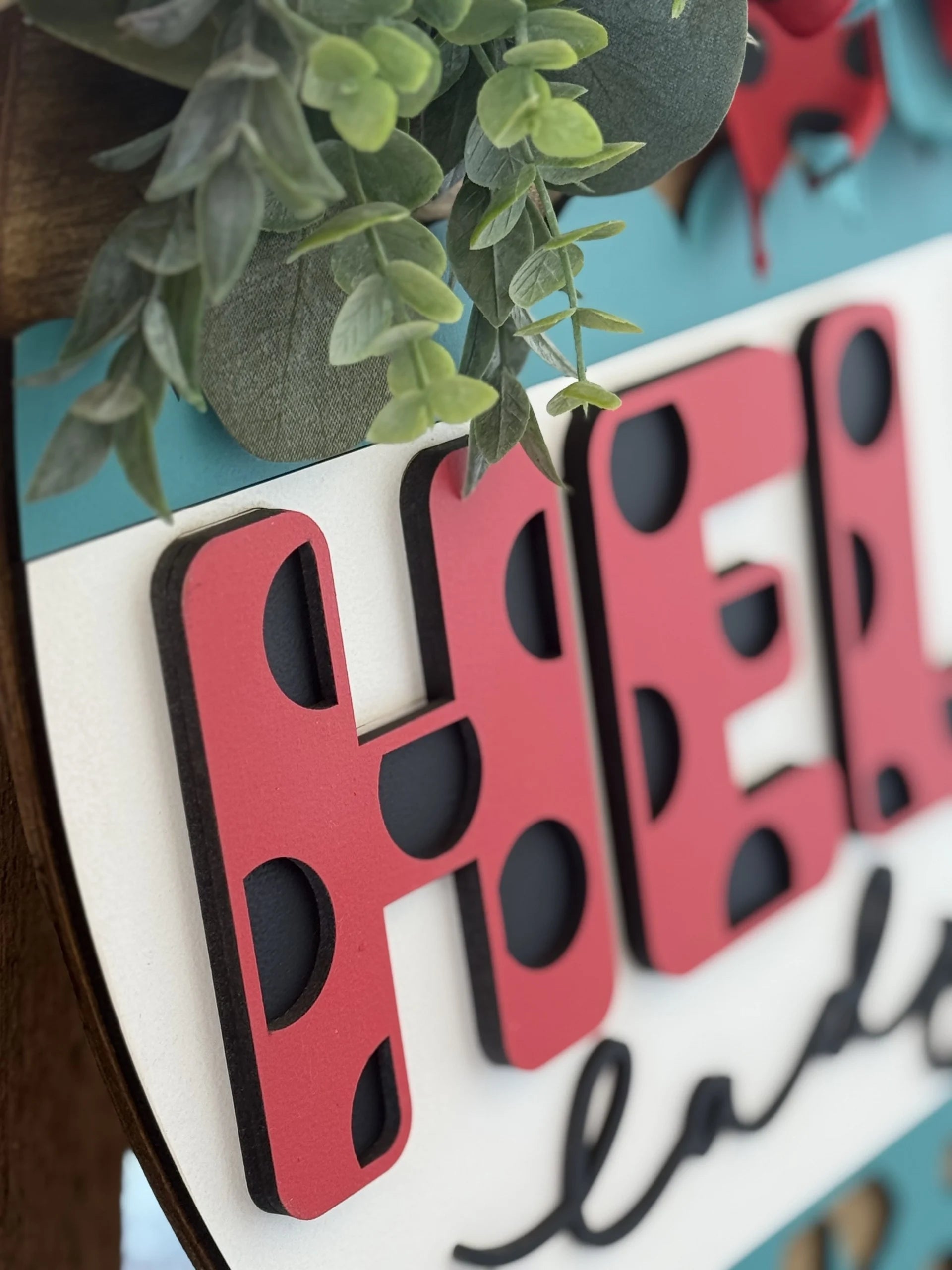 Close-up of a decorative sign with large, red letters reading HEL featuring black polka dots. Above the letters are green, leafy artificial plants. The sign has a blue background with portions of a red element visible at the top.
