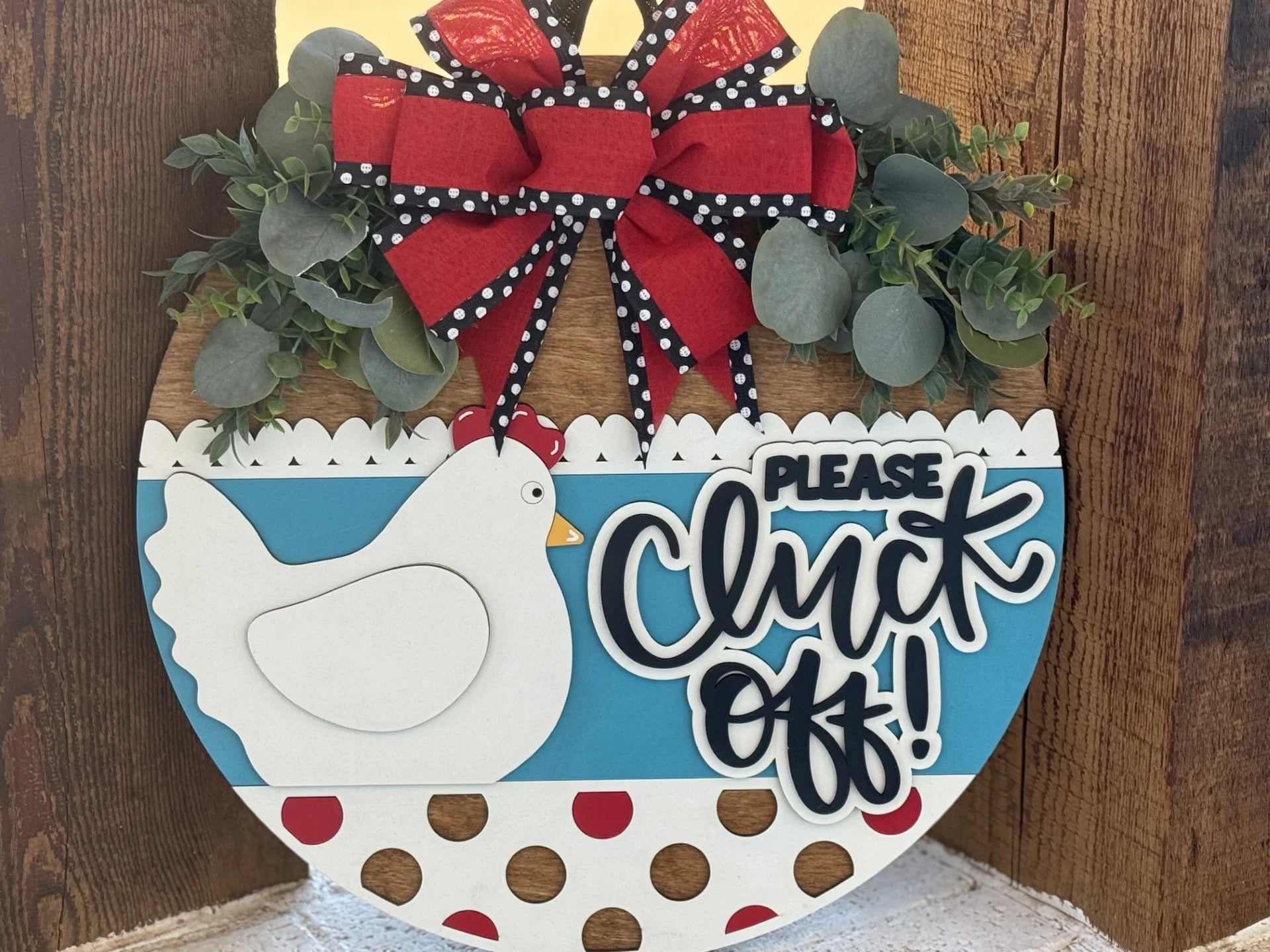 Round wooden sign with a decorative chicken, red and white polka dots, and blue background. It features a red and black polka-dotted bow, green foliage, and the phrase Please Cluck Off! in bold black letters. The sign is set against a rustic wood backdrop.