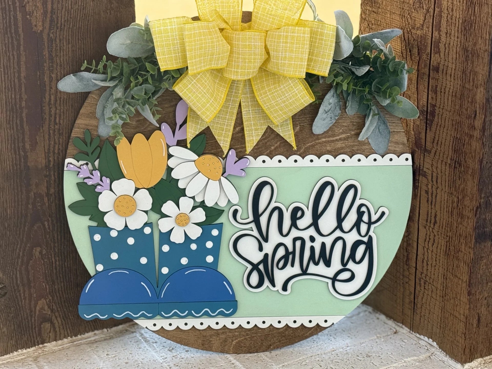 A round wooden sign features the text hello spring in elegant black script on a pastel green background. It is adorned with blue polka-dotted boots, spring flowers, and greenery. A large yellow gingham bow is attached at the top, adding a cheerful, seasonal touch.