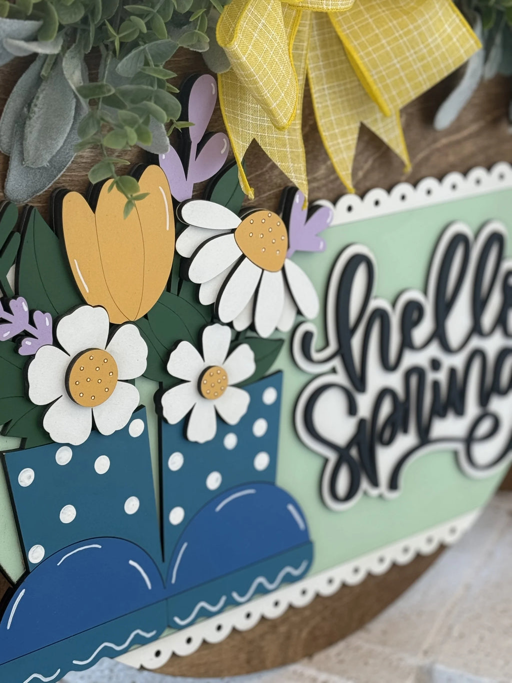Close-up of a decorative spring wreath. Features yellow tulips and white daisy-like flowers with green leaves in blue polka dot rain boots. A yellow ribbon is tied at the top. The words hello spring are in bold script on a mint green background with a scalloped white border.